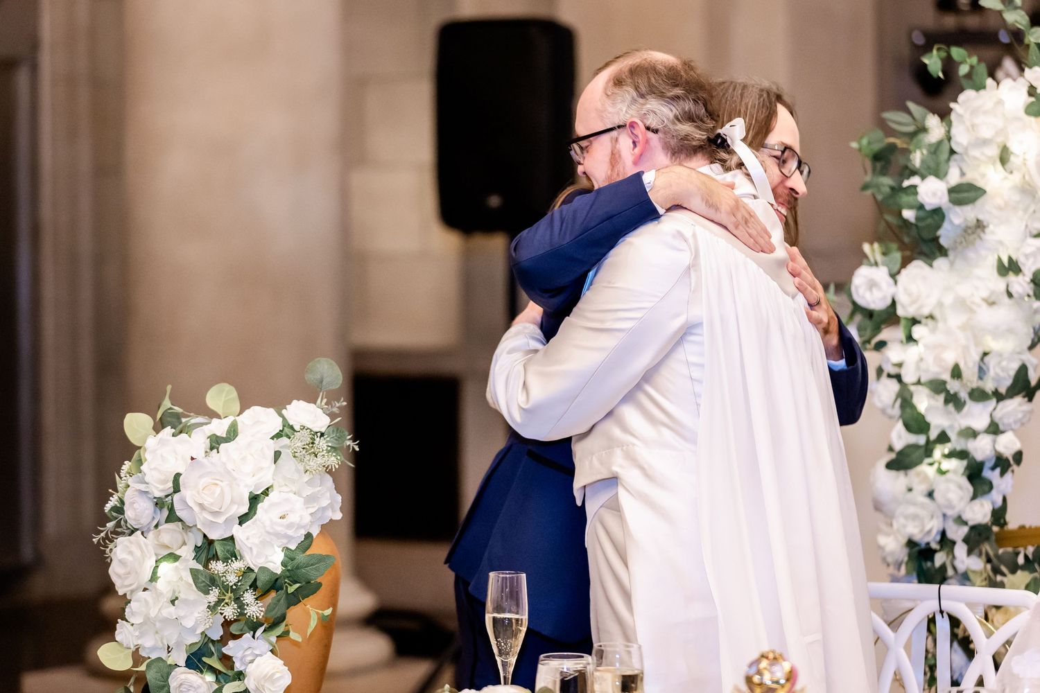Emotional embrace shared during wedding reception under elegant floral arrangements.