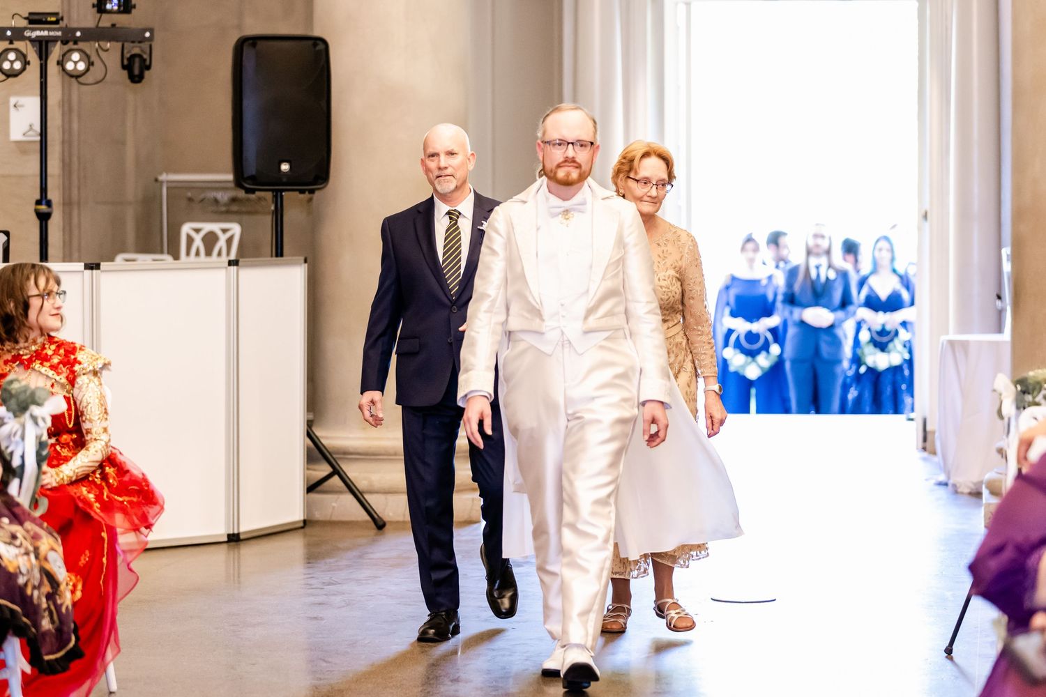 Wedding processional in a bright venue with formal attire and decorative red accents.