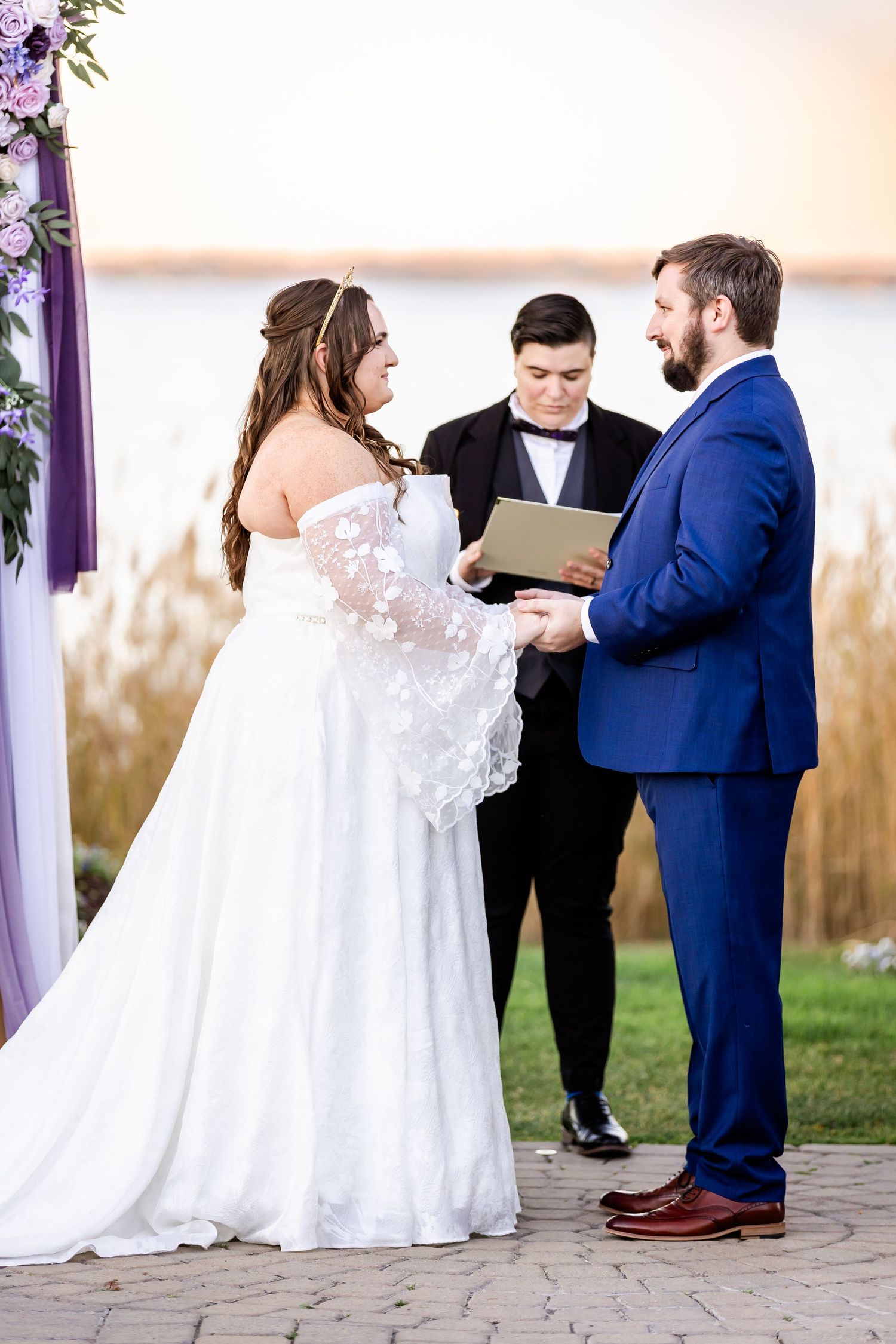 Wedding ceremony vows being exchanged under decorated arch with waterfront backdrop.