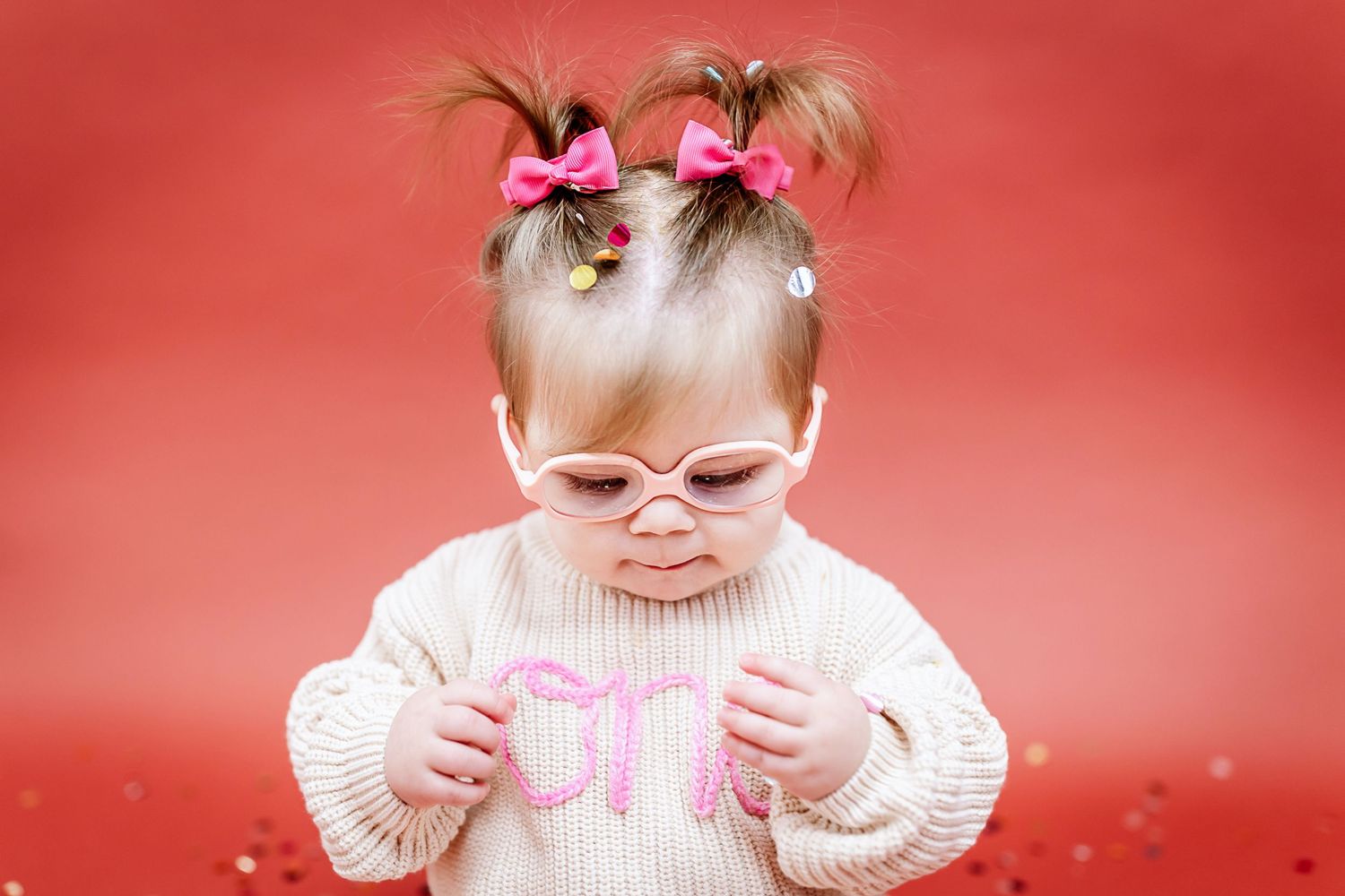 Sweet baby in glasses and pink sweater sitting on red background.