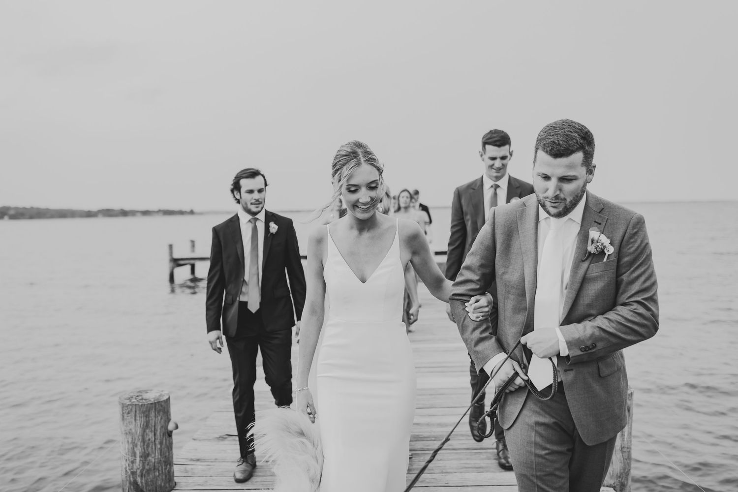A couple in wedding attire walk along a waterfront dock with groomsmen following behind in black and white.