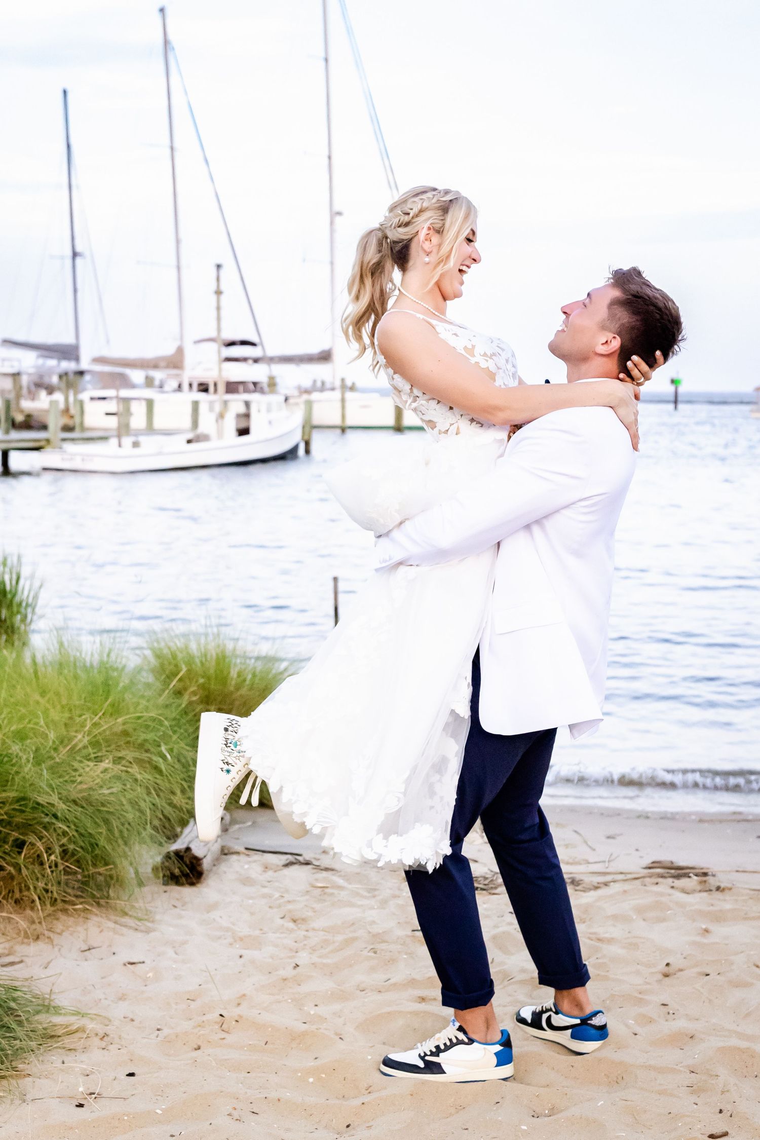 A couple celebrates at a marina with boats in background as he lifts her in white dress while wearing blue sneakers.
