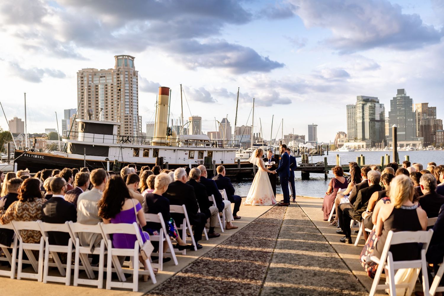 Wedding ceremony on a waterfront dock with city skyline backdrop and crowd seated in white chairs.
