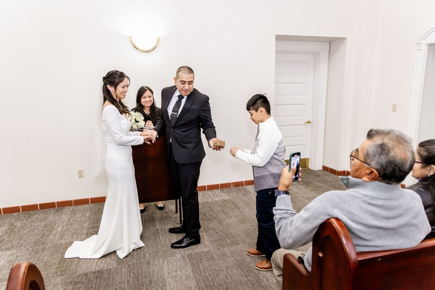 Wedding ceremony in progress with guests watching from wooden benches in a simple indoor setting.
