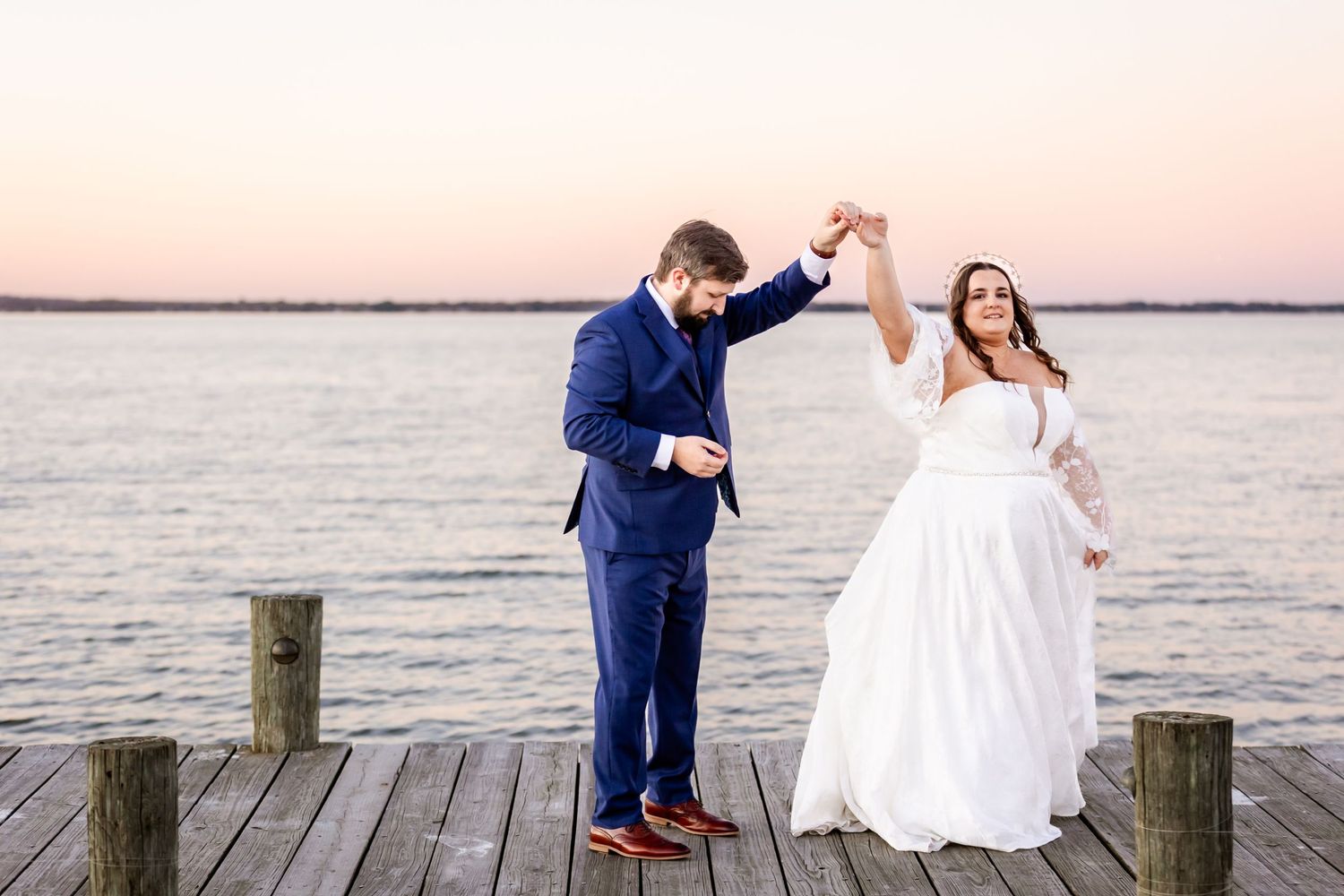 A couple dances together on a wooden pier at sunset during their wedding celebration.