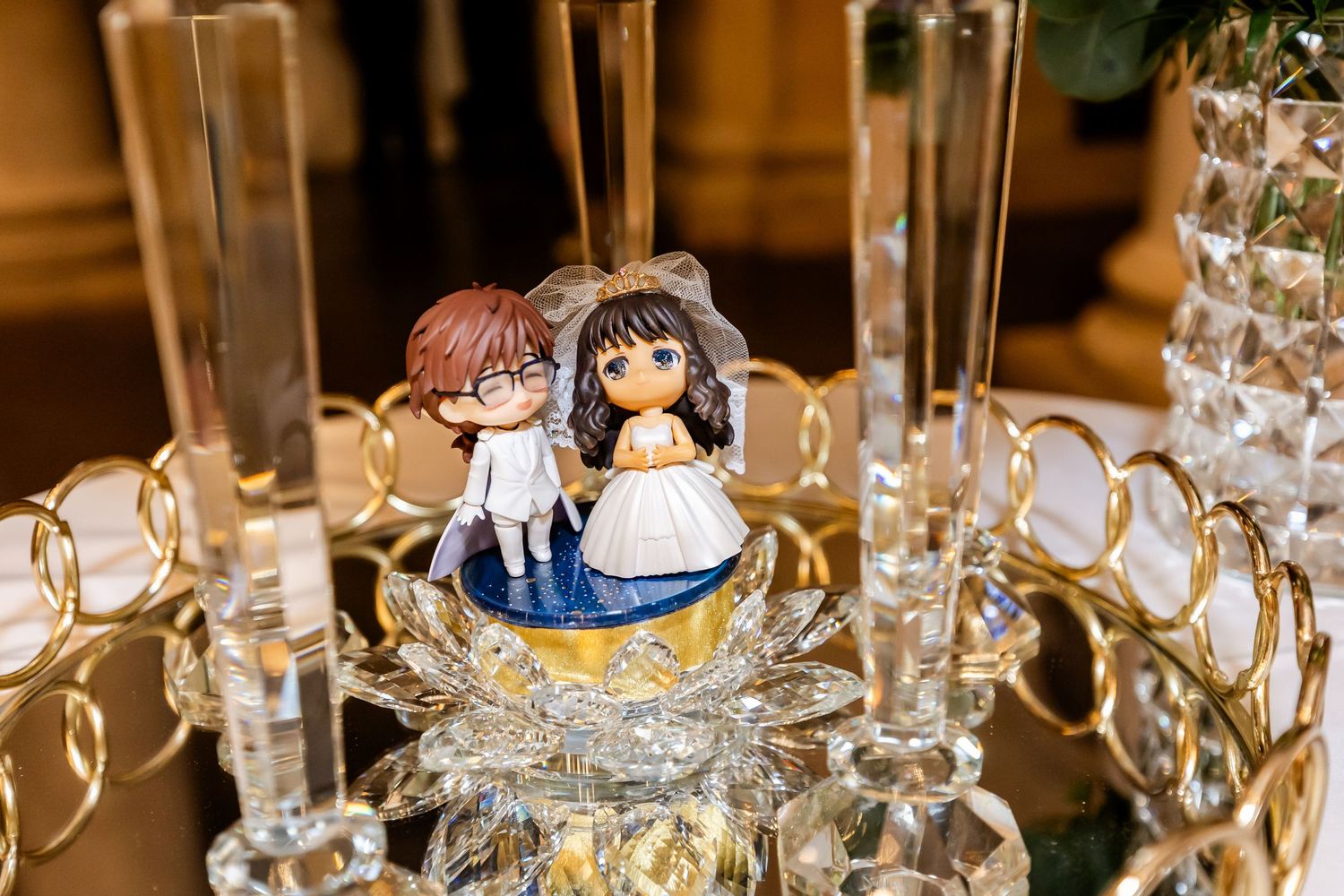 Cute anime-style cake topper figurines displayed on crystal stand at wedding reception.
