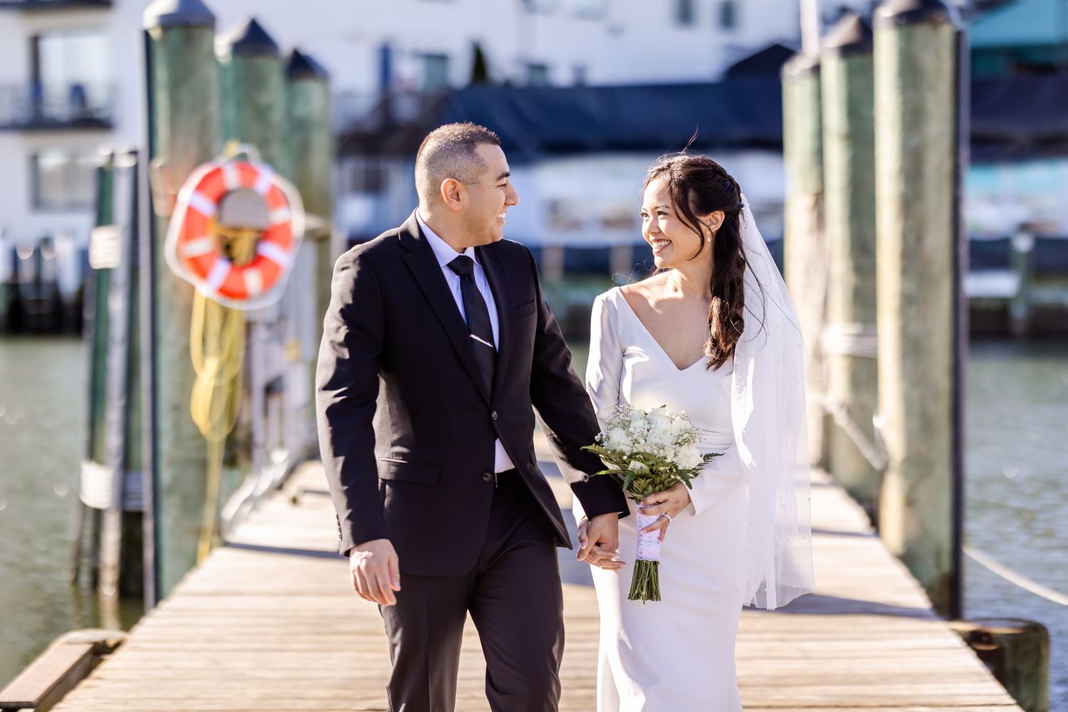 Wedding couple stands on a wooden pier overlooking the water on a sunny day.