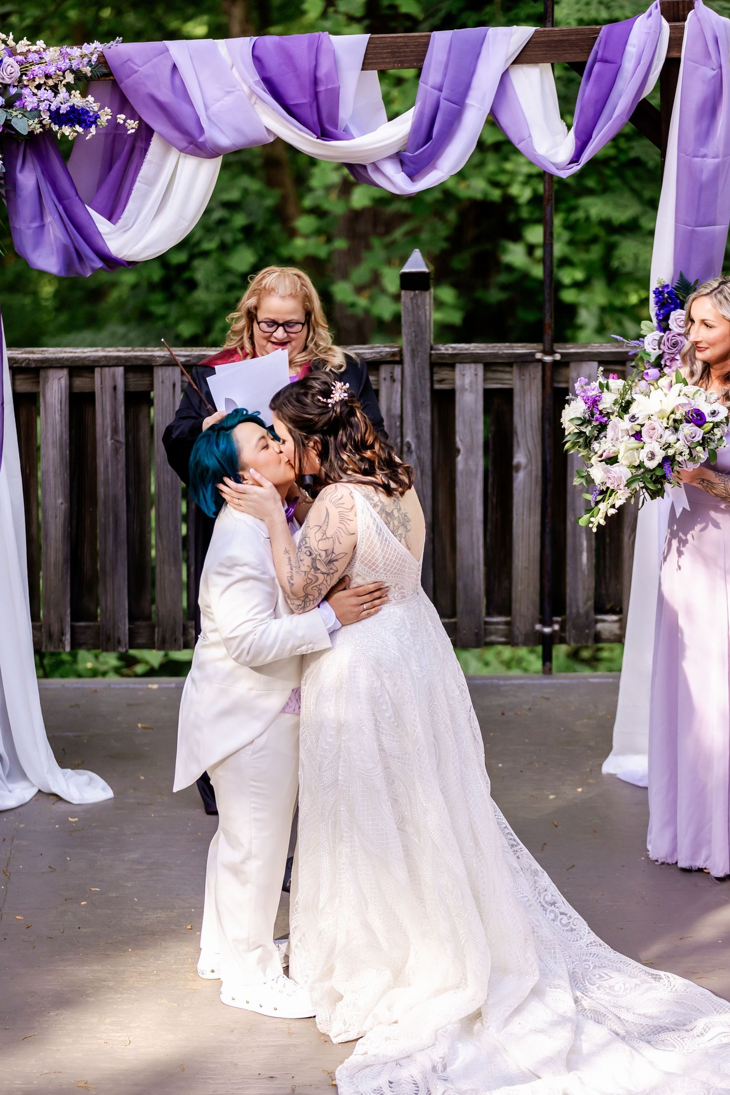 A romantic first kiss at a wedding ceremony beneath purple and white draped decorations.