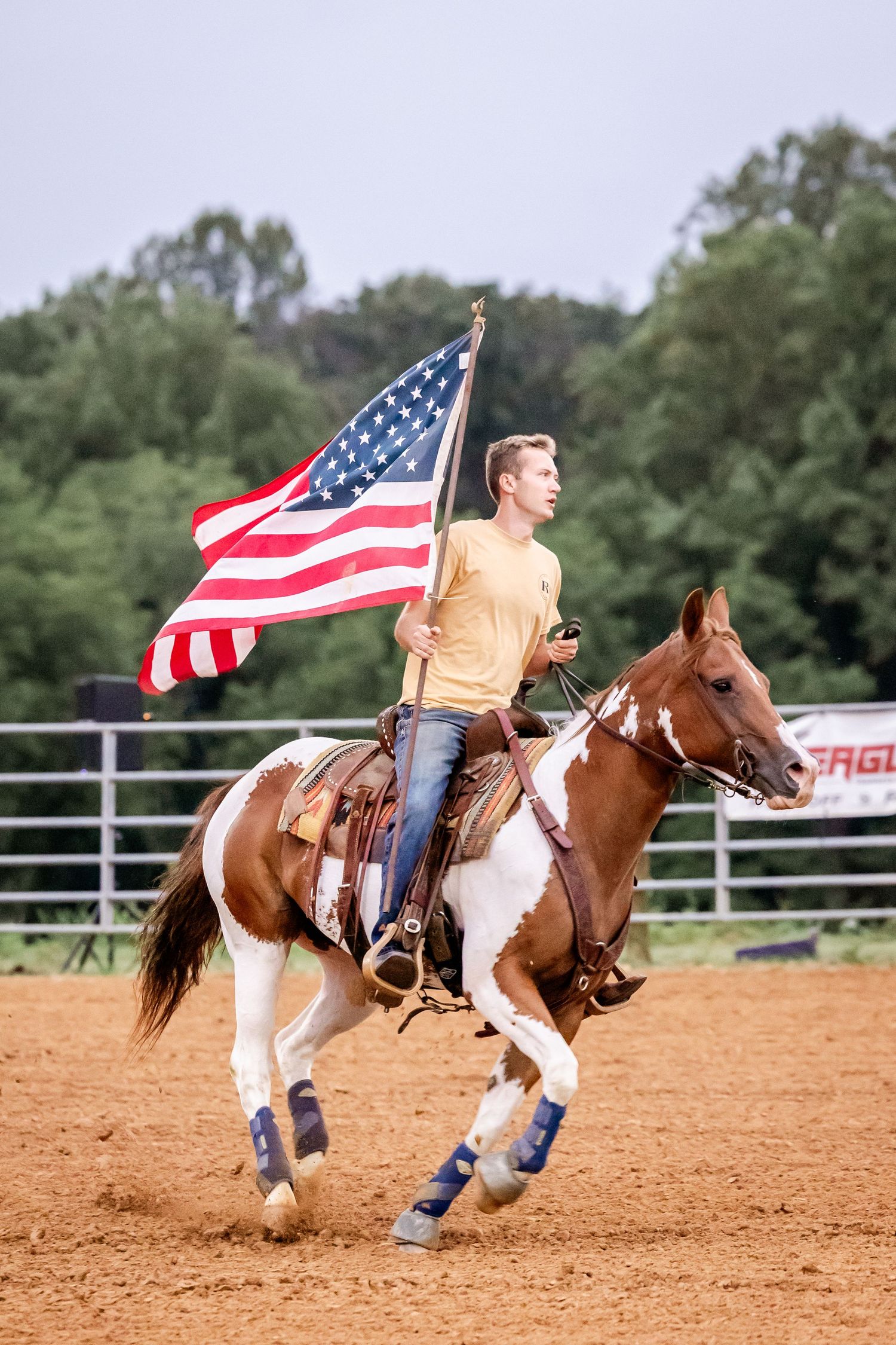 Rider carrying American flag while galloping on paint horse in rodeo arena.