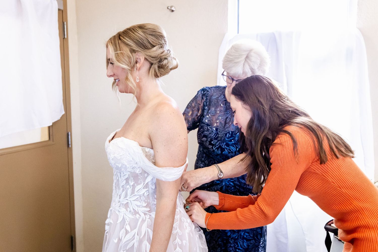 Wedding preparations show helpers assisting with a white lace bridal gown in a sunlit room.