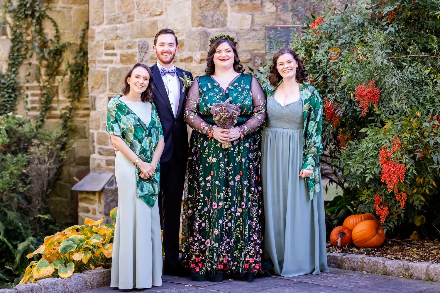 A group portrait in formal attire stands against a stone wall with autumn decorations and pumpkins.