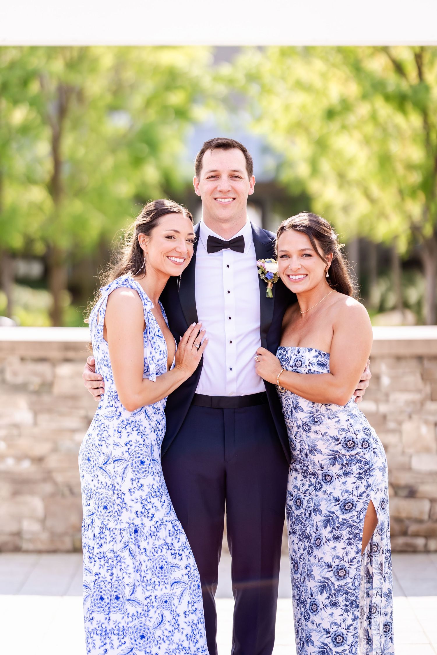 Wedding party members in blue floral dresses and black tuxedo posing outdoors.