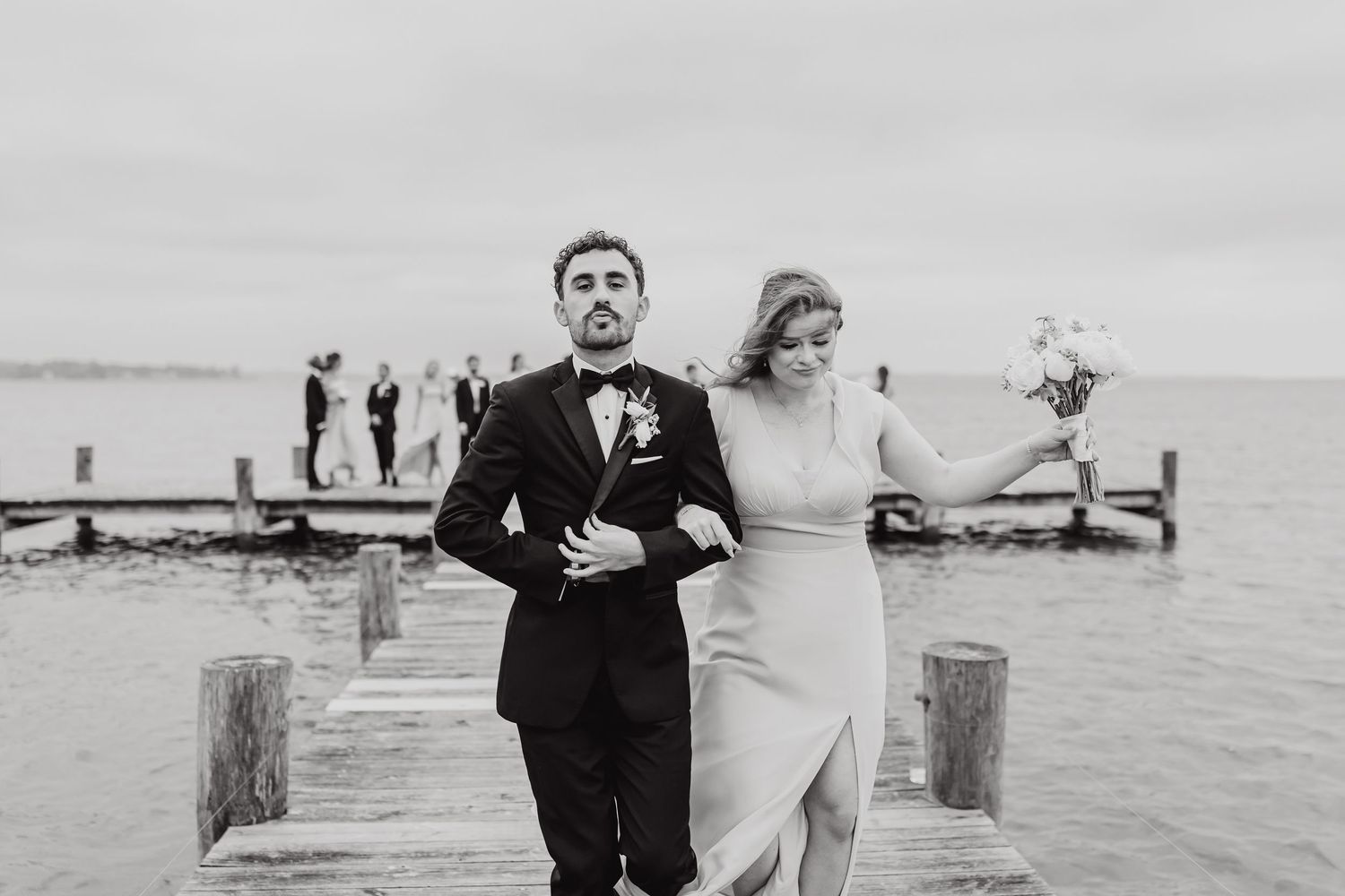 A black and white photo of a couple walking along a wooden dock by the water.