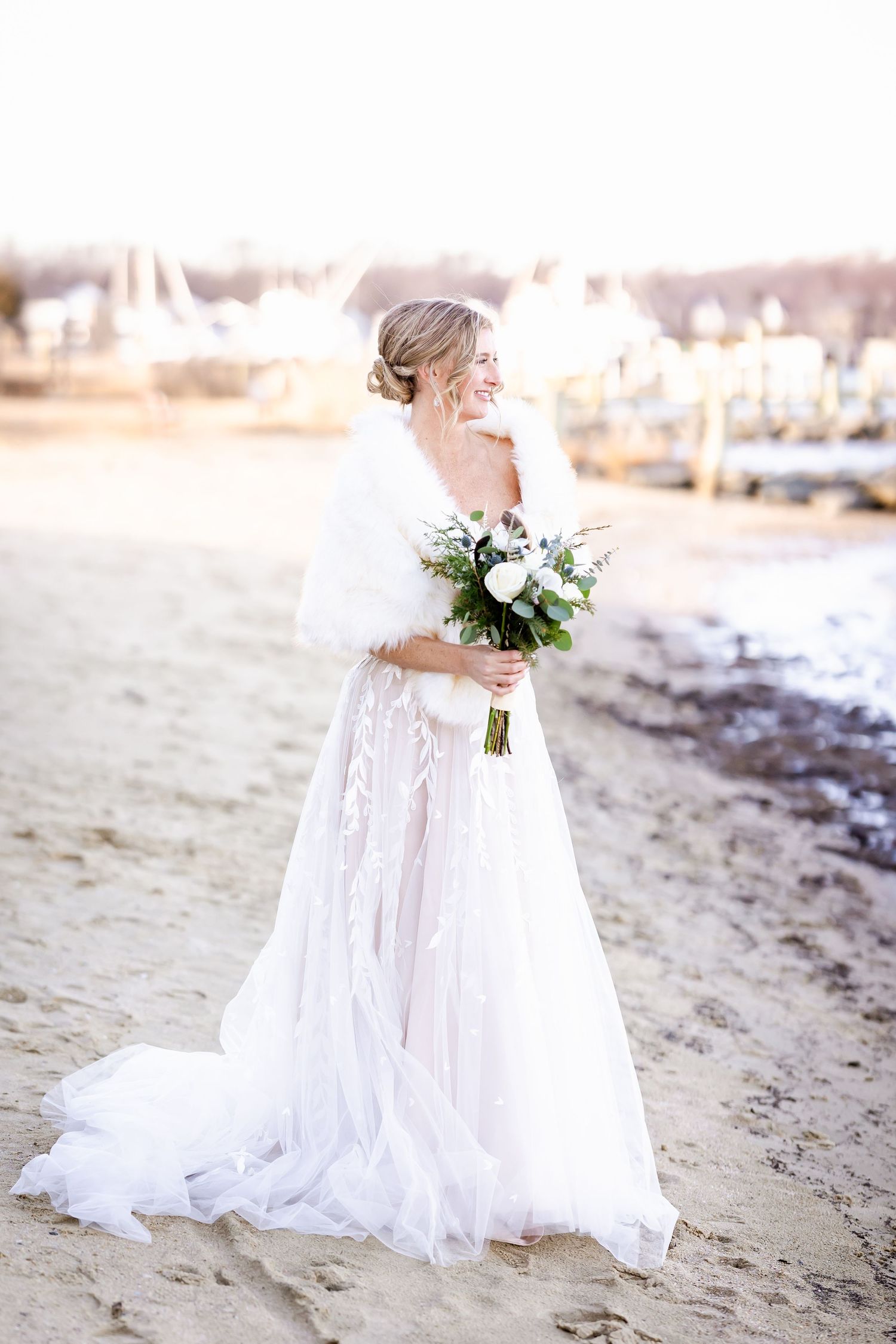 Elegant bride in flowing white gown holds white floral bouquet while standing on beach at sunset.