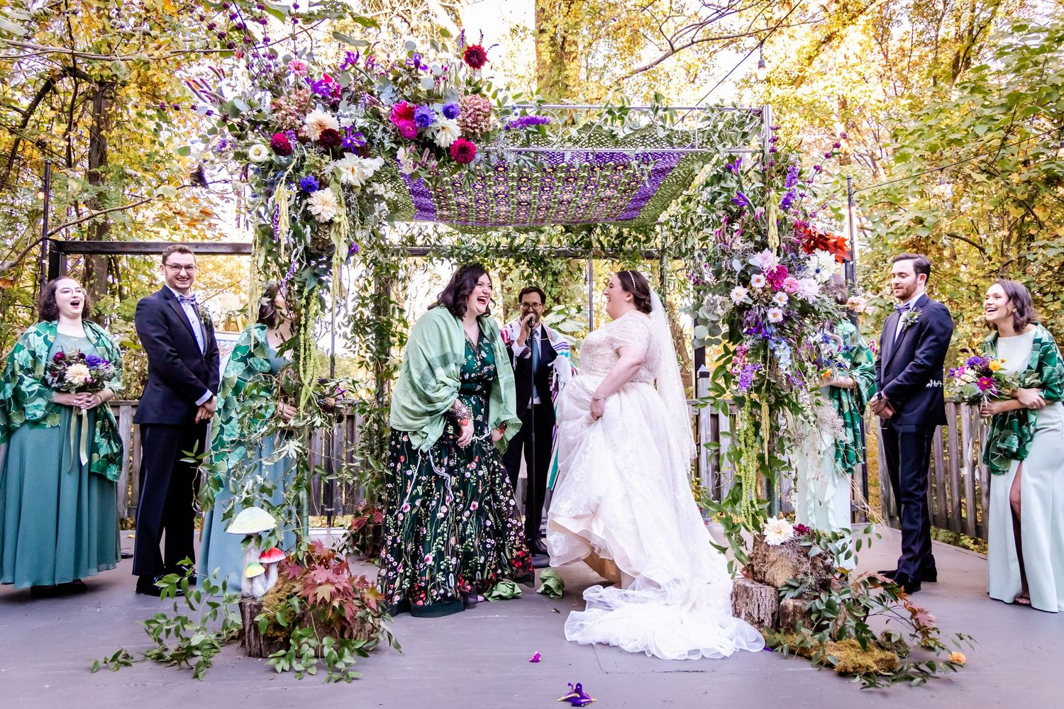 Wedding ceremony setup with purple floral arch and wedding party under trees.