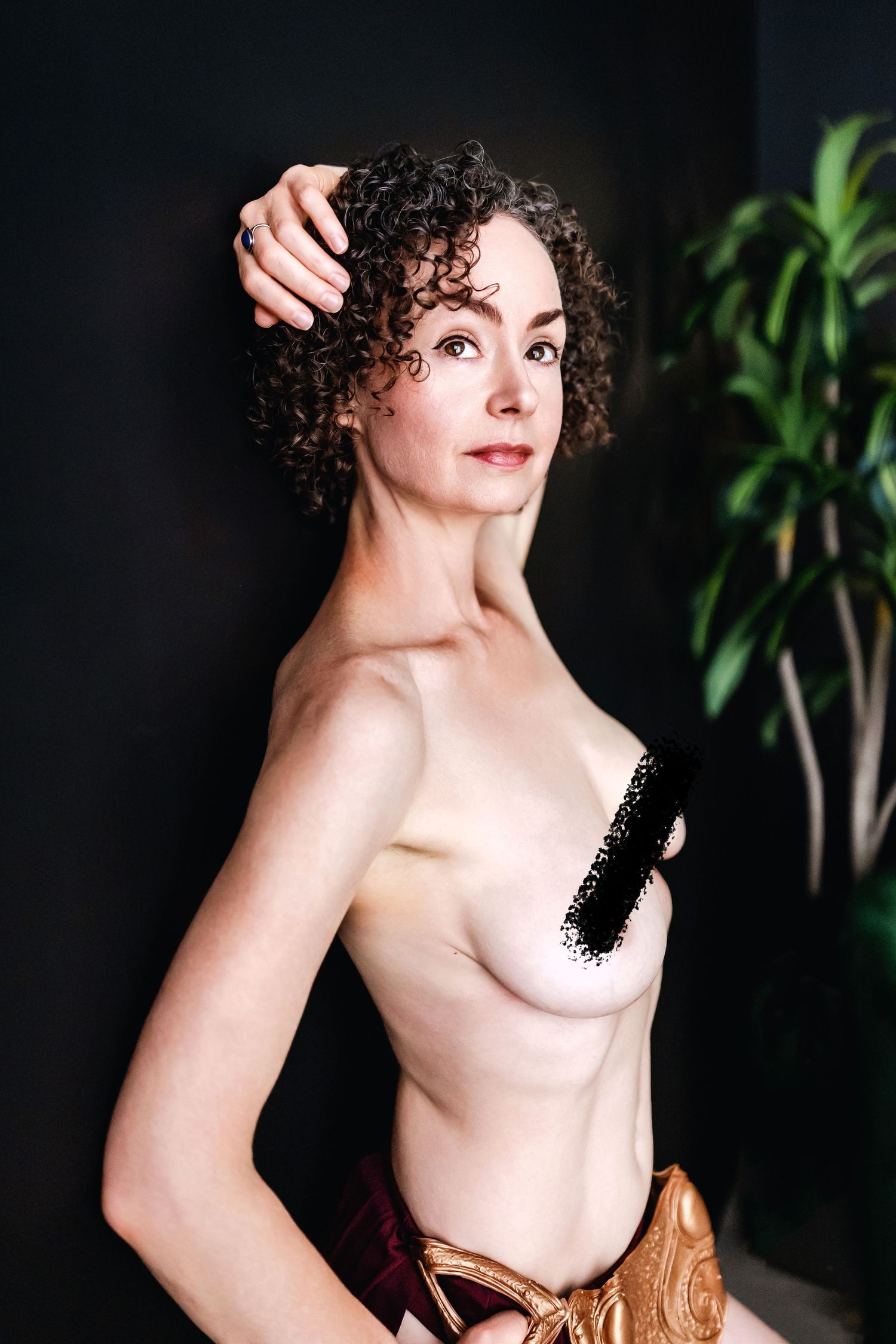 A person with curly dark hair poses in an artistic portrait with a partial black top against a dark background.