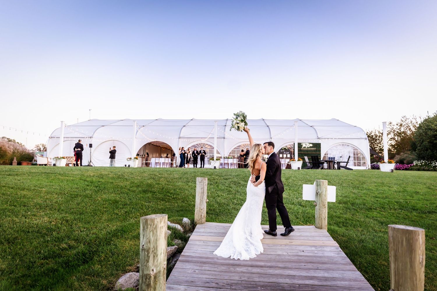 A wedding couple stands on a wooden dock with a white reception tent and horses in the background.