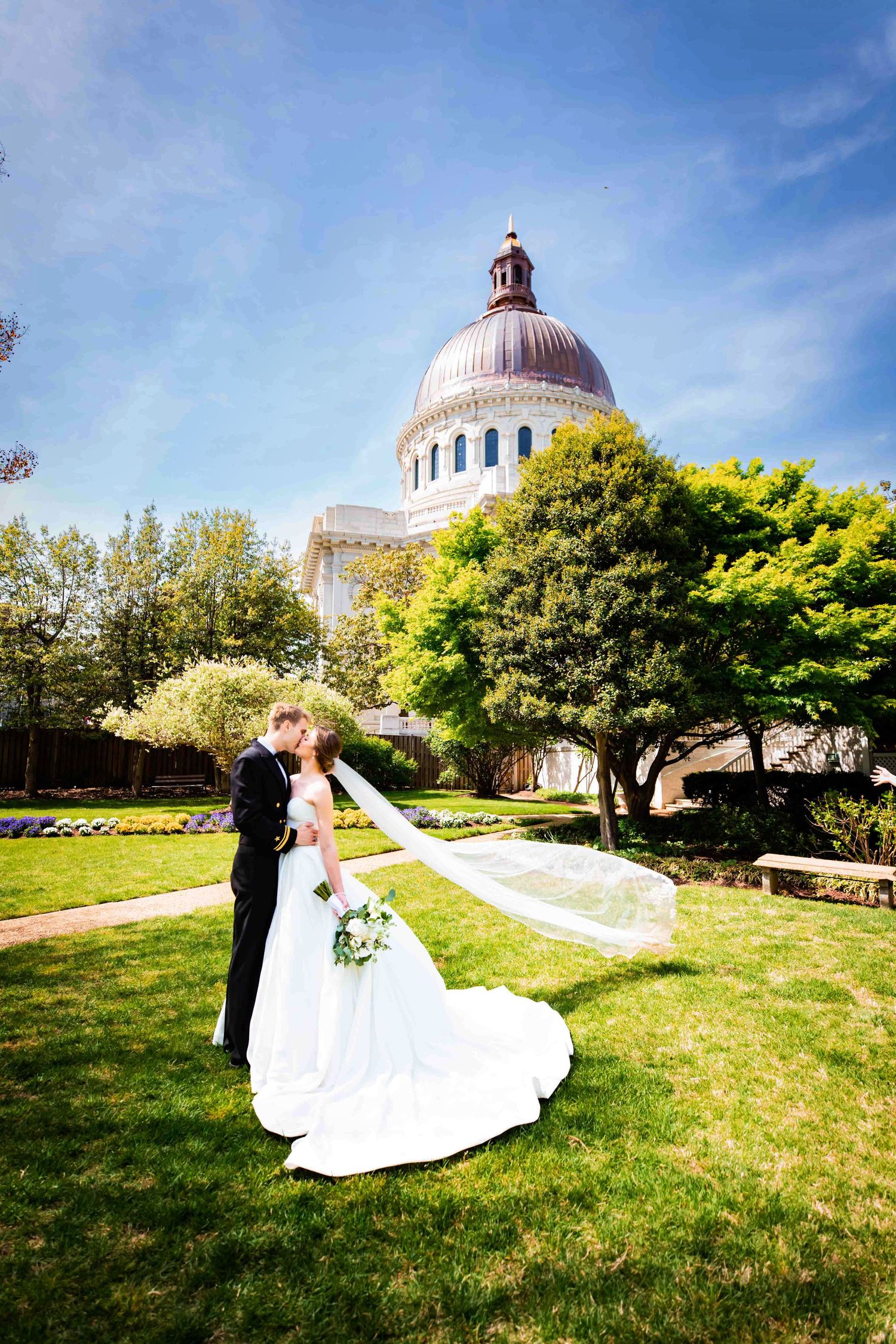 A couple in wedding attire share a romantic moment on a lawn with a grand domed building in the background.