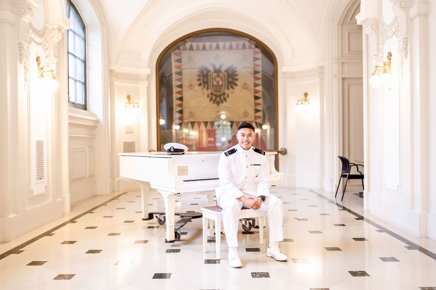 Figure in white naval uniform sits at grand piano in elegant marble hallway with arched ceiling and ornate decor.