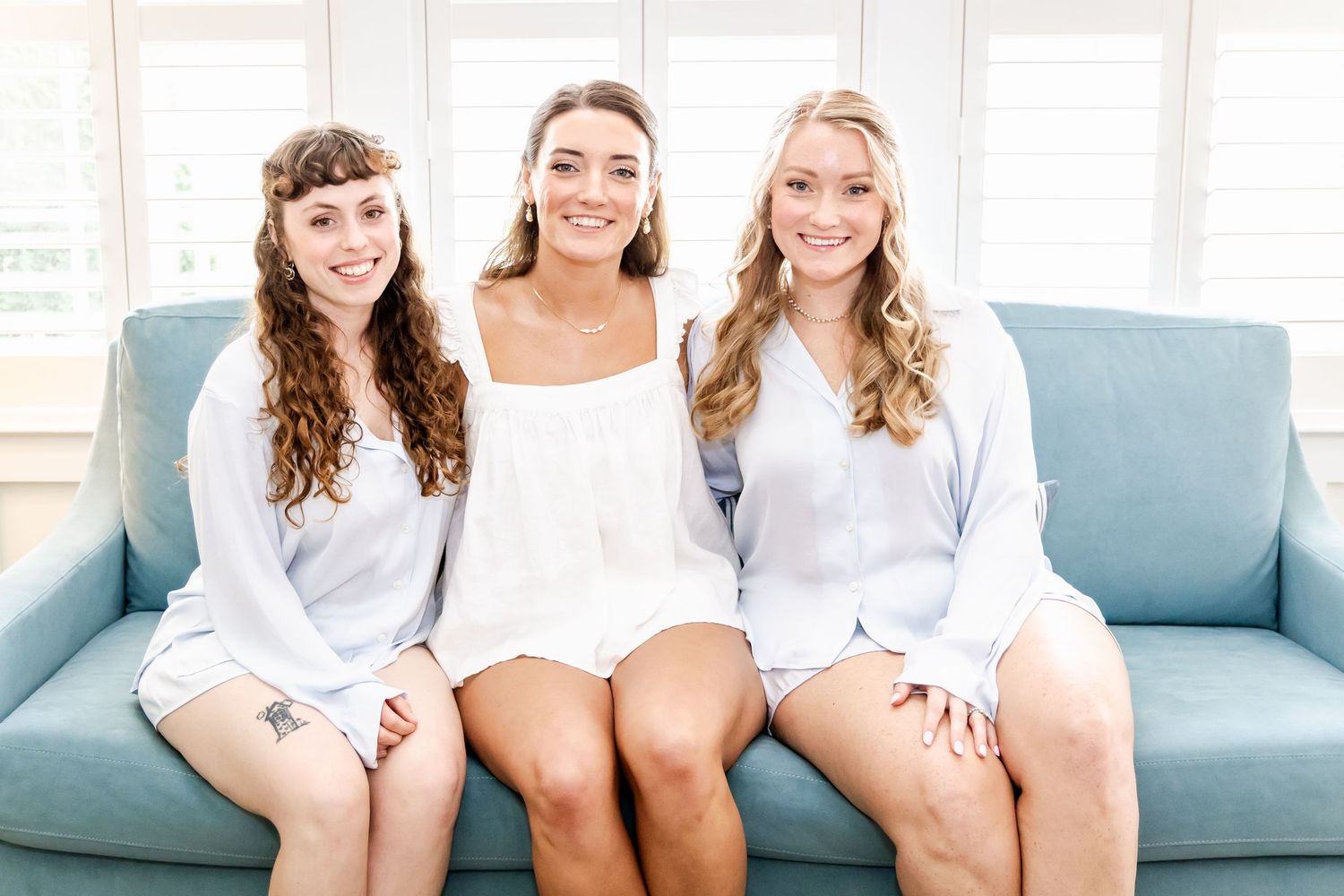 Bridesmaids in cozy white robes sitting together on light blue couch during wedding preparations.