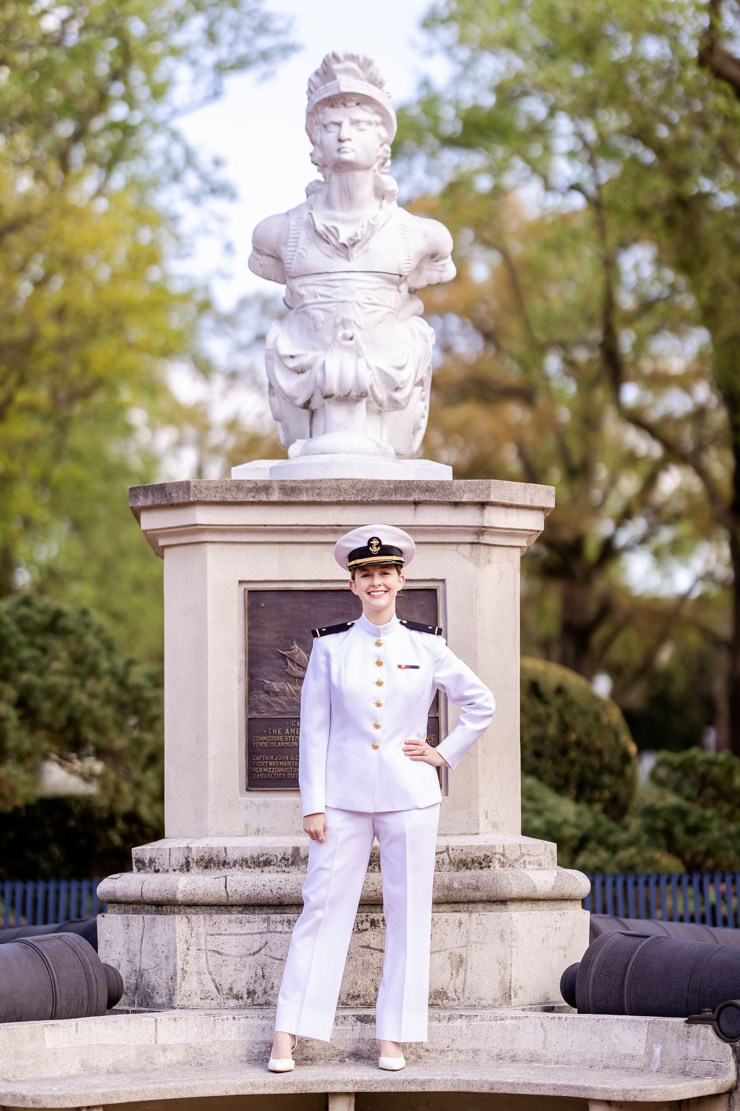 A naval officer in dress whites stands at attention in front of a classical stone monument and bust in a garden setting.