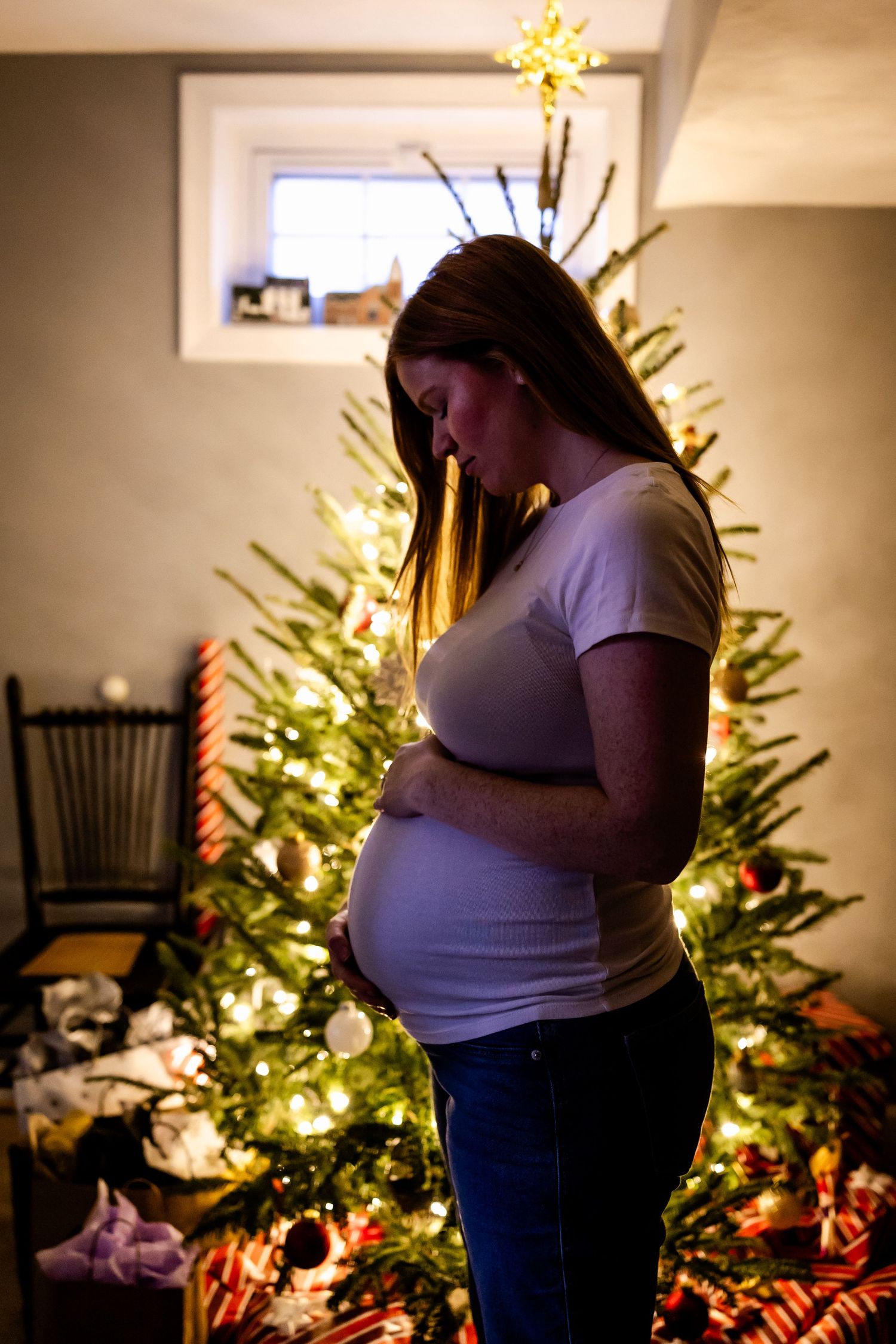 A silhouetted expectant mother stands in front of a glowing Christmas tree decorated with lights and ornaments.