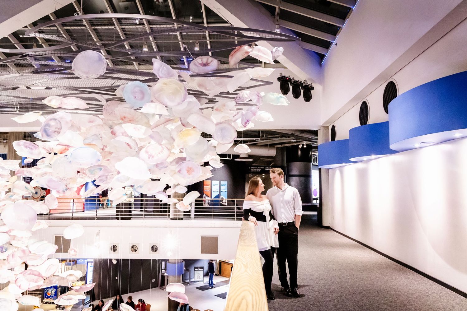 People stand beneath a modern art installation of suspended white objects in a museum space.