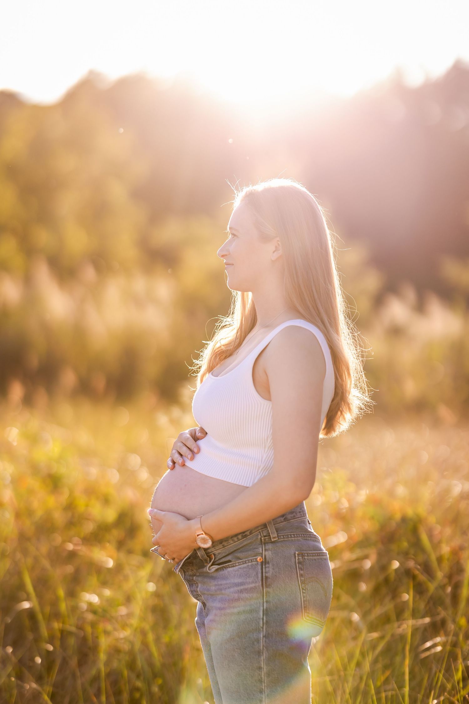 Pregnant woman in white tank top and jeans standing in golden sunlit field.