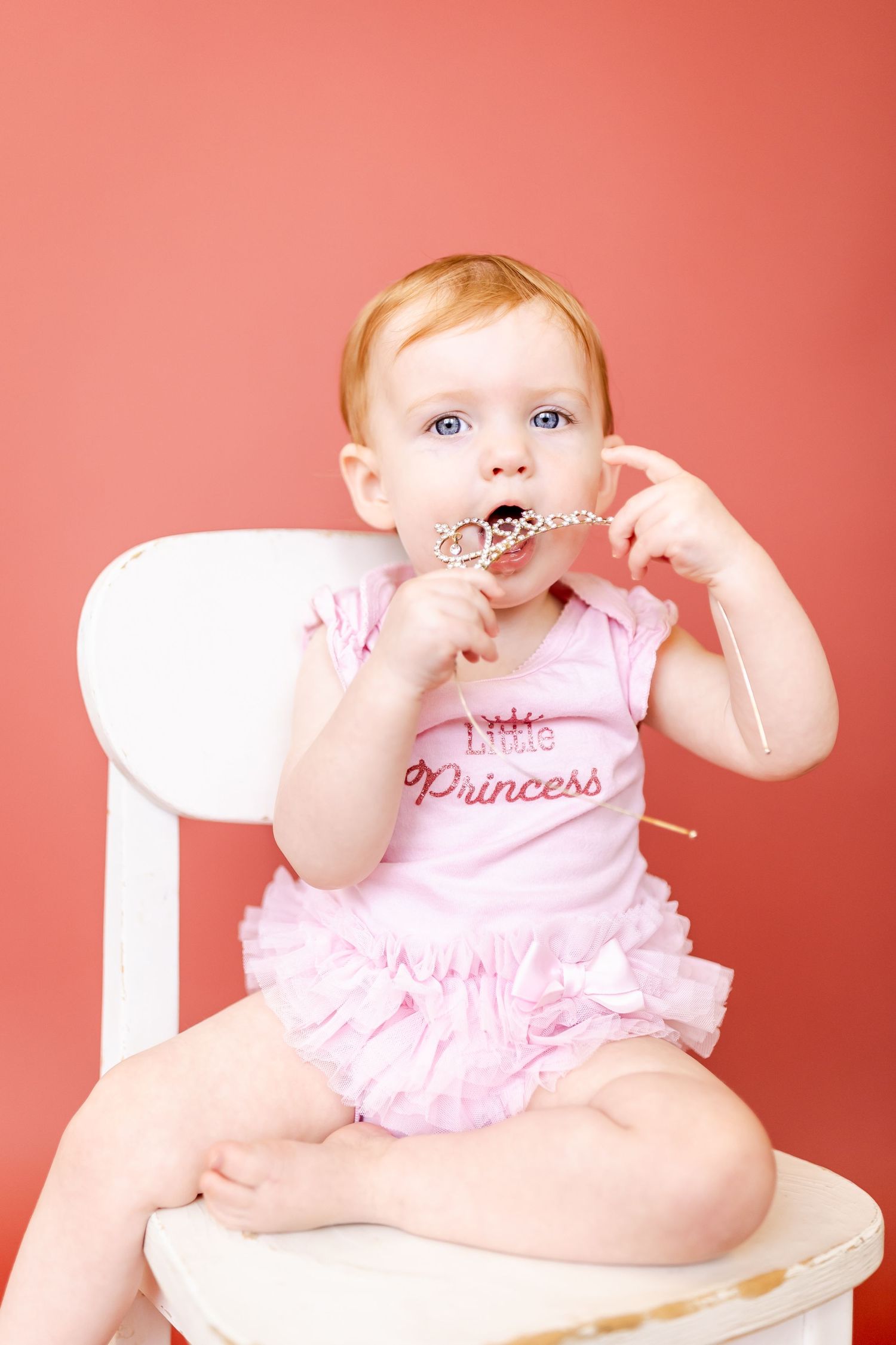 Baby wearing princess outfit poses in white chair against pink backdrop.
