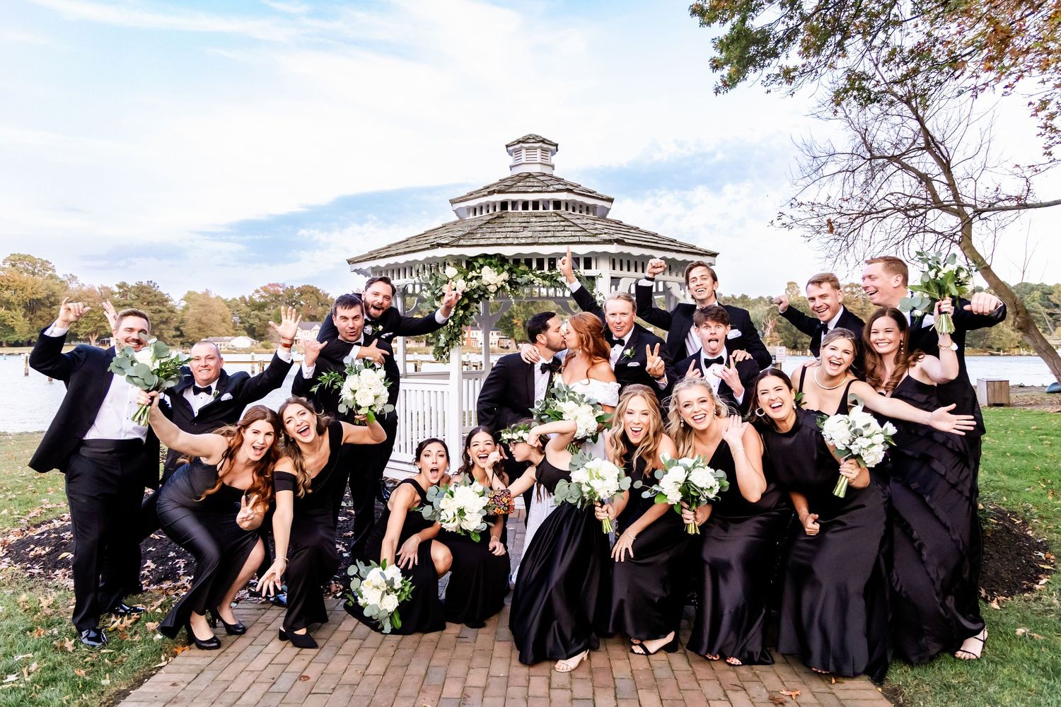 Wedding party in black attire poses playfully for photos on a sunny day near a gazebo and waterfront.