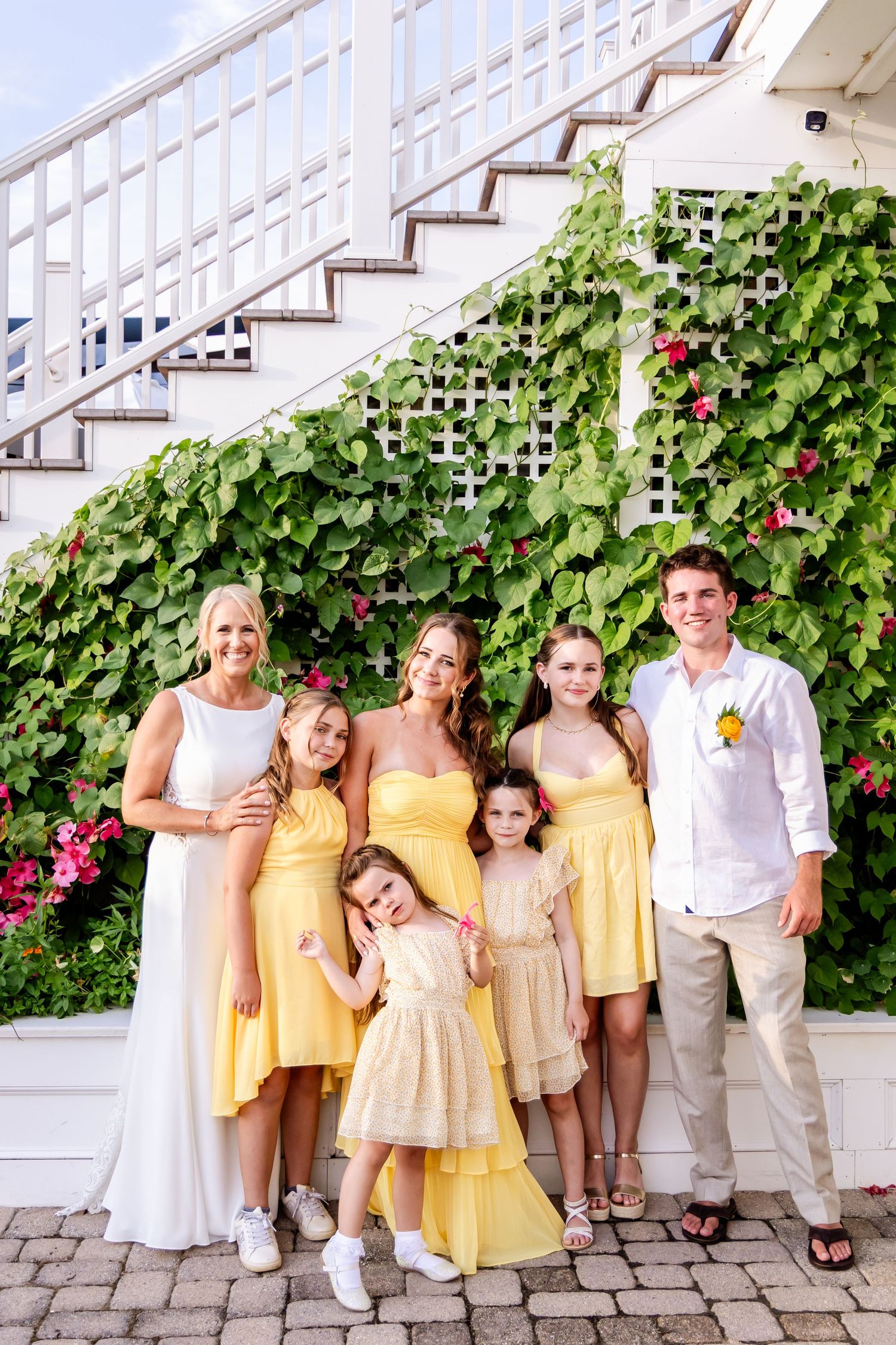 A group in coordinated yellow and white outfits poses together on stone steps with lush green vines and pink flowers.
