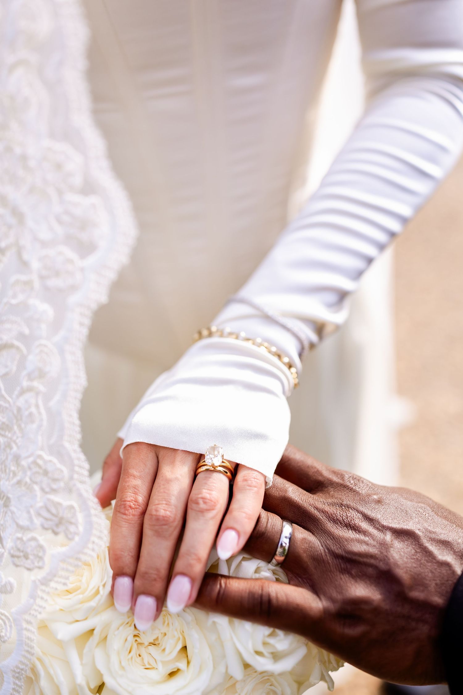 Bride and groom hands together showing wedding rings, bride wearing white gloves holding bouquet of white roses.