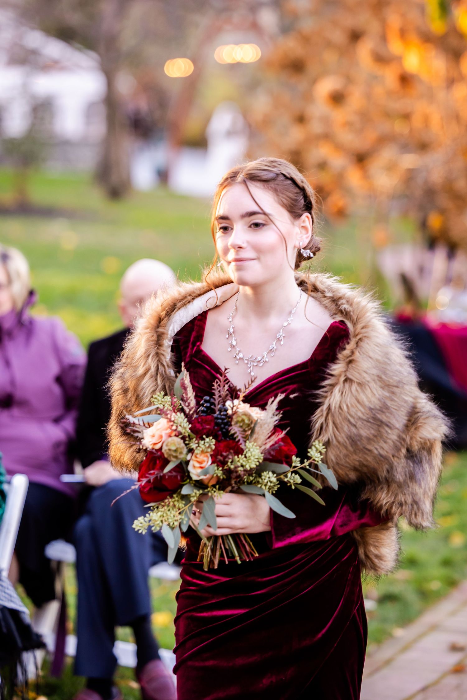 Bride wearing burgundy velvet dress with fur stole and floral bouquet in autumn setting.