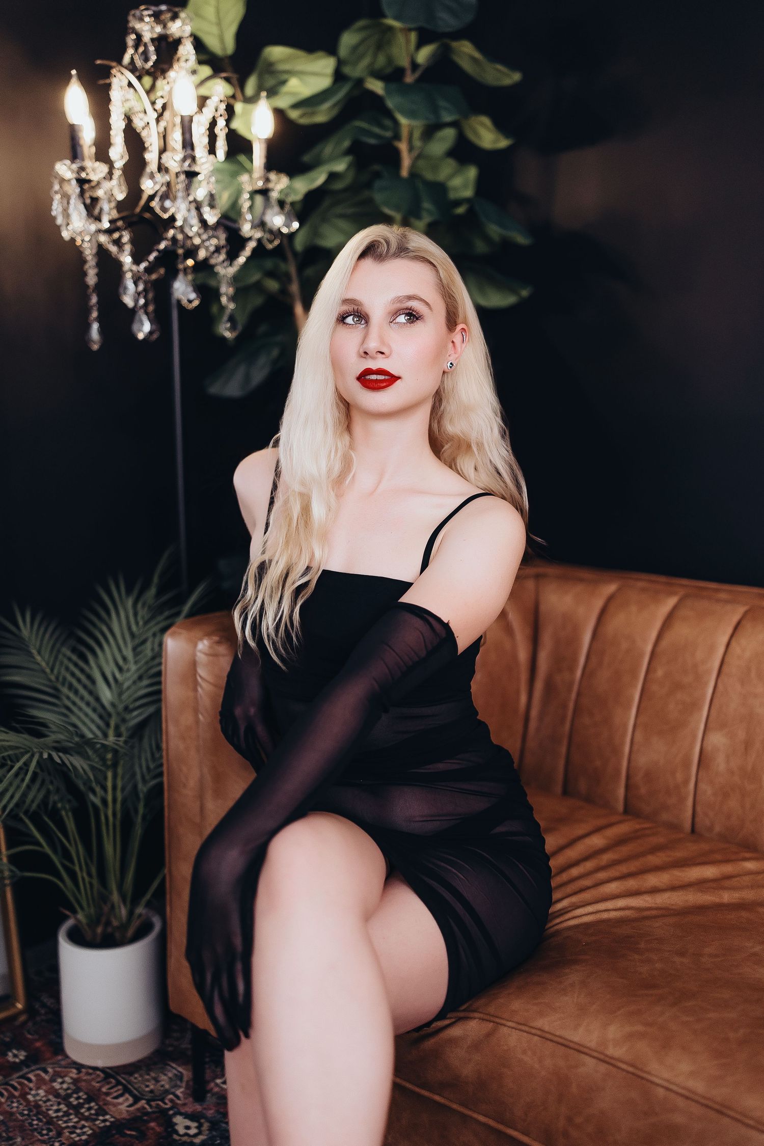 A glamorous boudoir photoshoot featuring a model in black attire posing on a brown leather couch under a crystal chandelier.