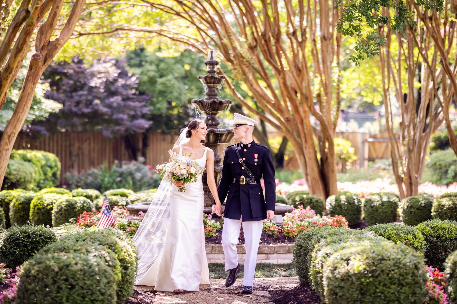 A military wedding couple walks through a garden path lined with crepe myrtle trees.