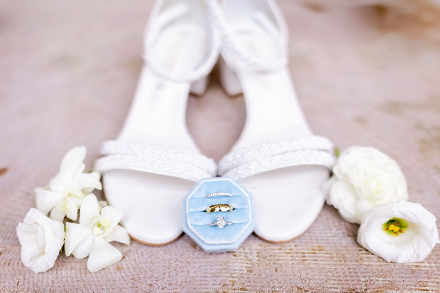 White bridal shoes with a blue button and white flowers arranged on burlap fabric for wedding details photography.