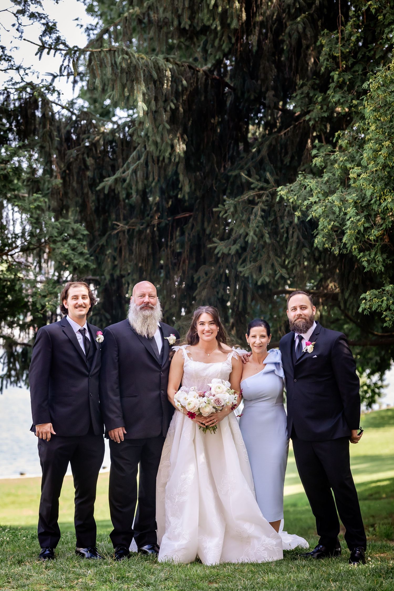 Wedding party in formal attire posing for group photos outdoors under large trees.