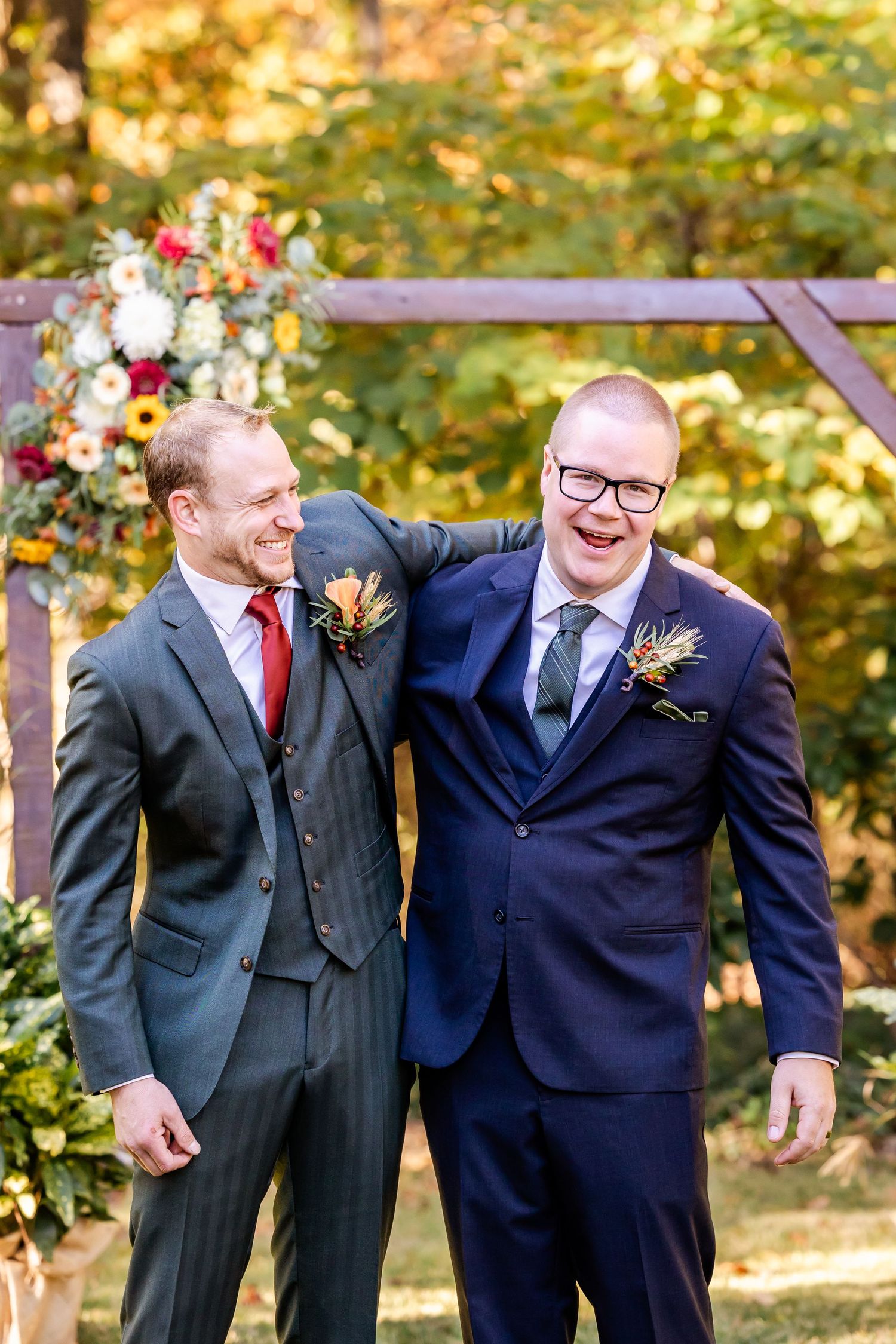 Two groomsmen in suits share a laugh under a floral wedding arch in an autumn setting.