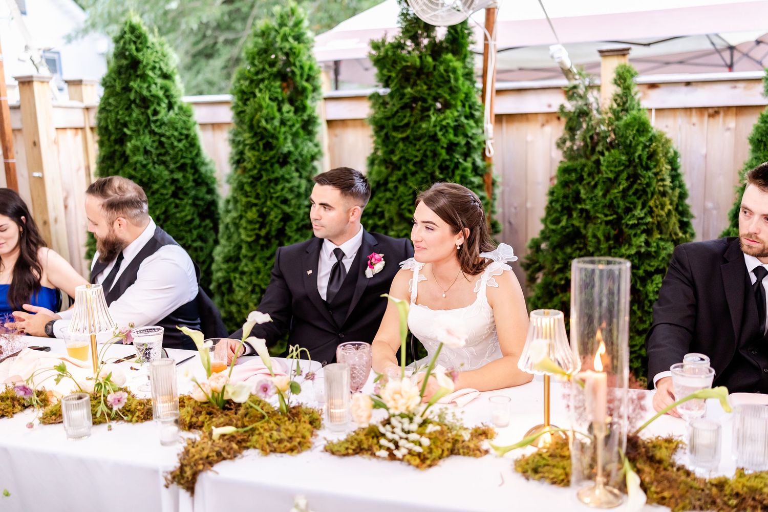 Long wedding reception table with guests dining under a white tent surrounded by greenery.