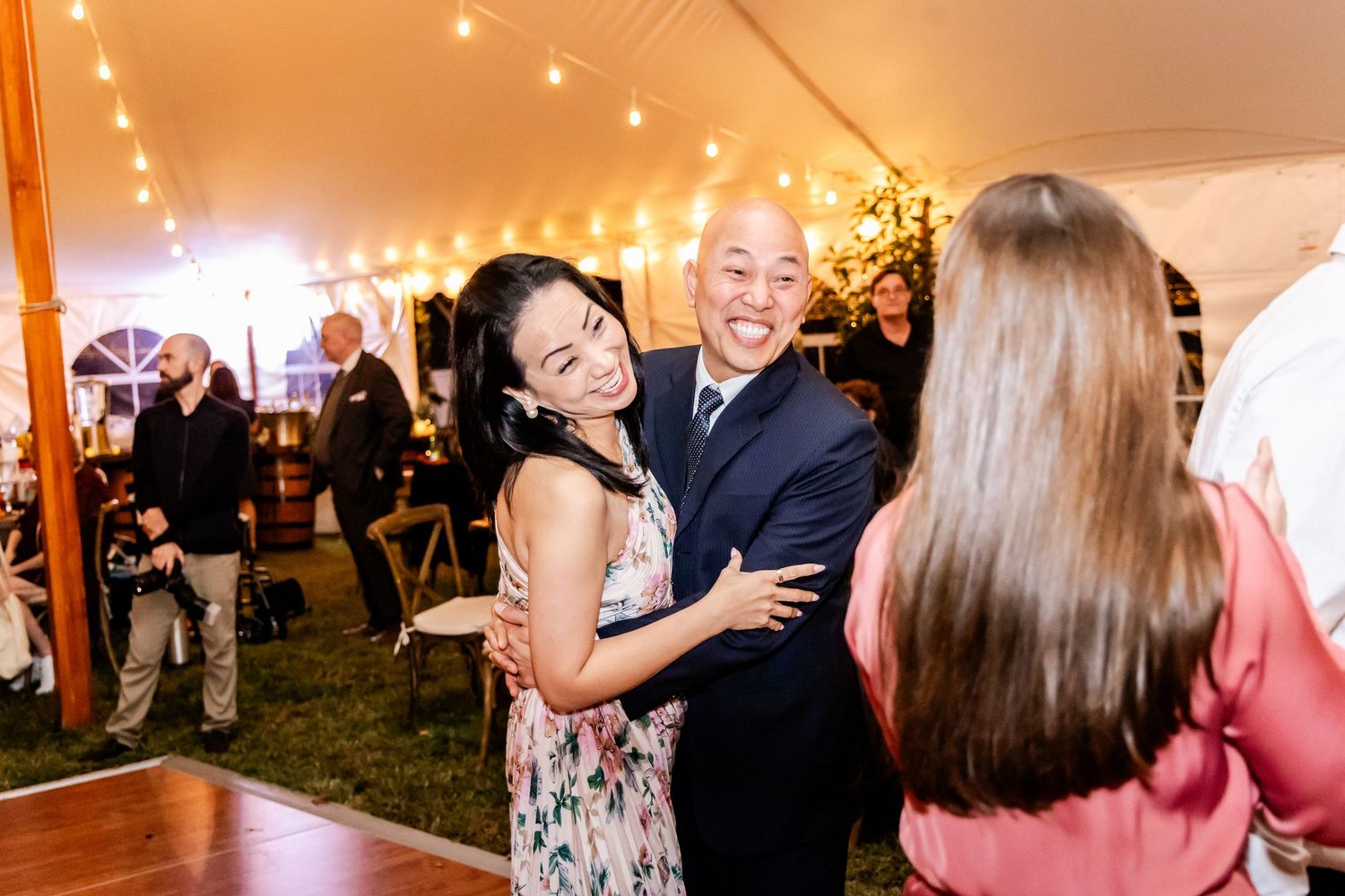 Wedding guests dance and celebrate under warm string lights in an elegant reception tent.