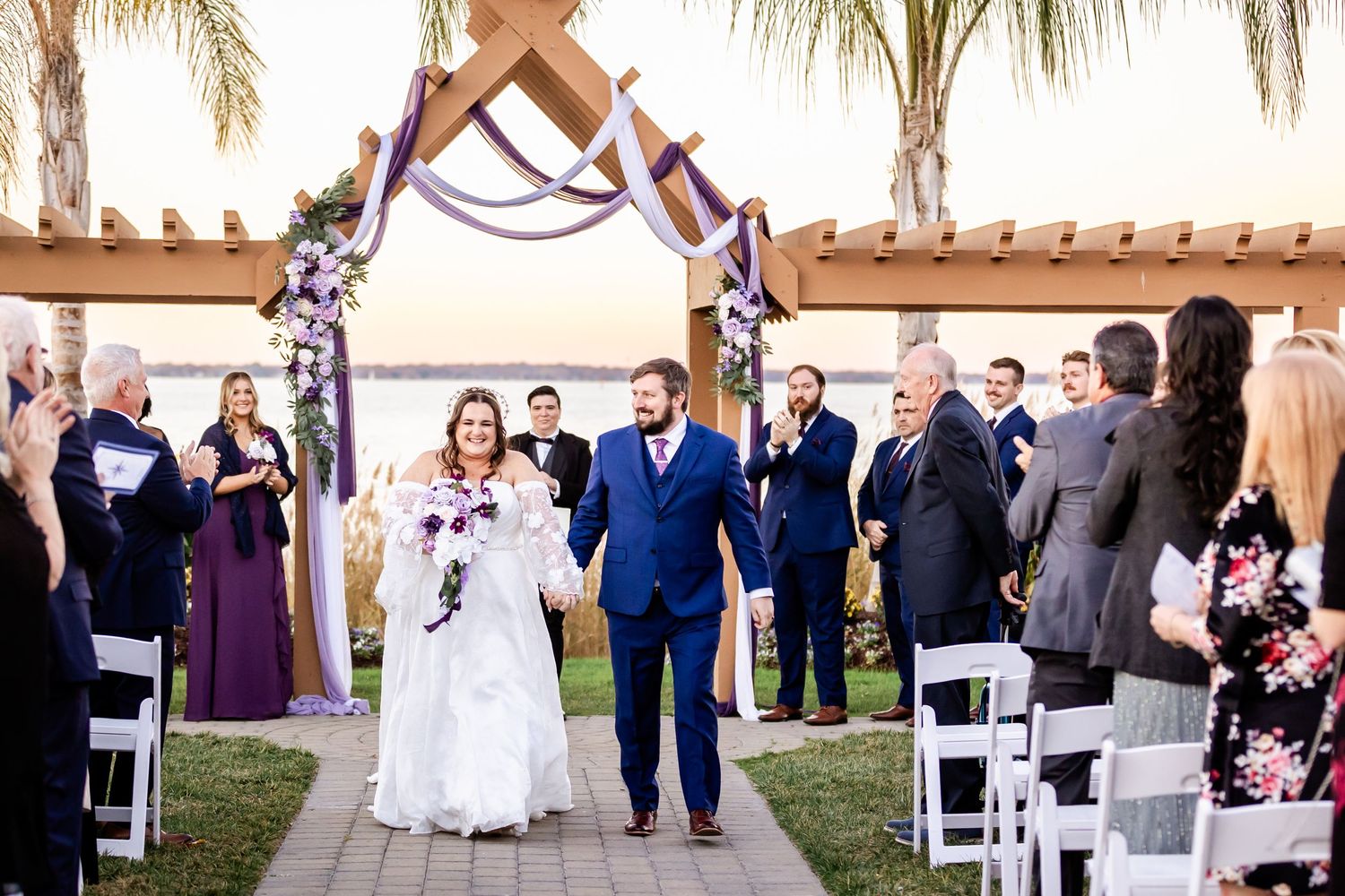 Newlyweds walk down the aisle under a decorated arch with guests standing by.