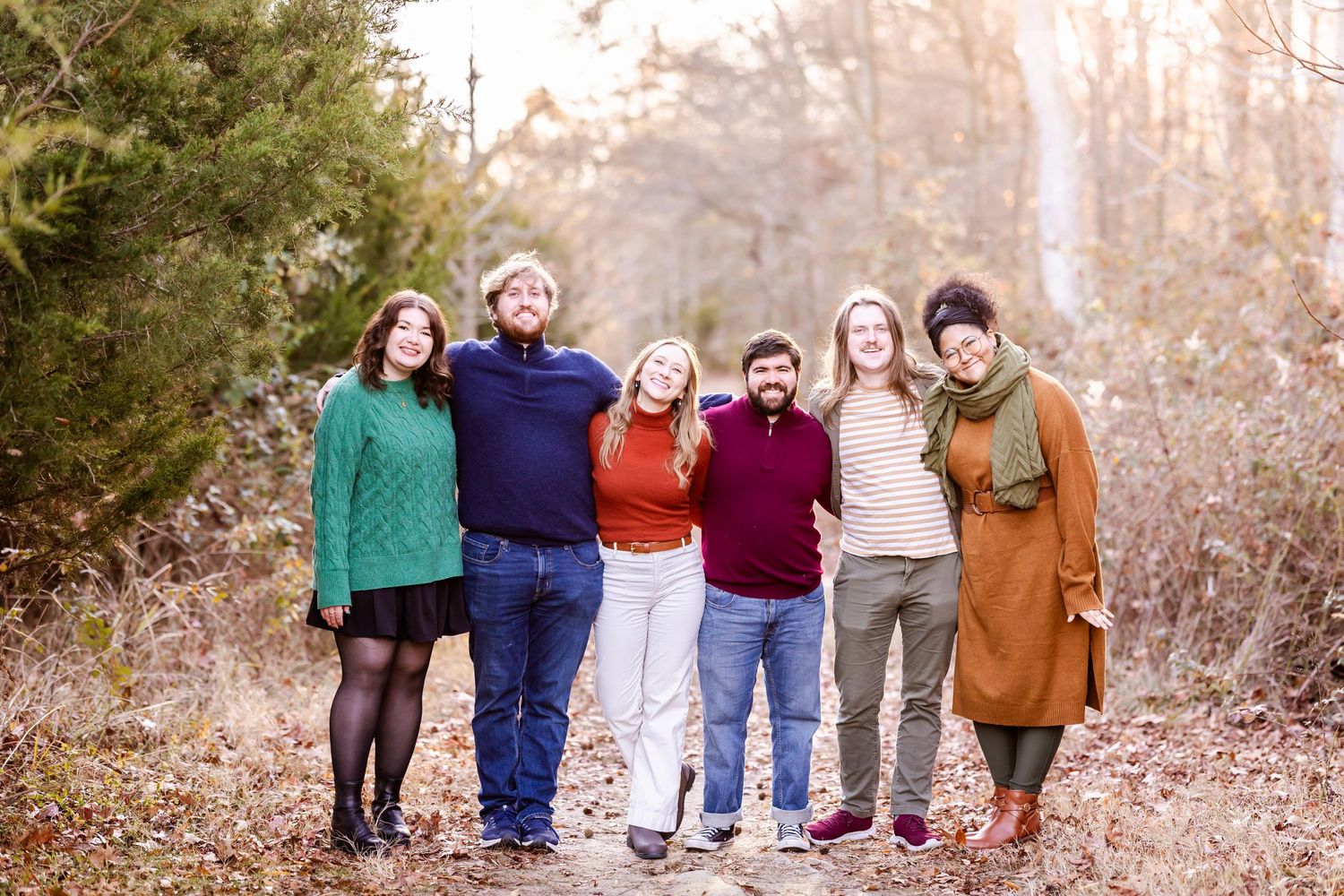 A cheerful group photo of six people standing together on a leaf-covered forest trail.