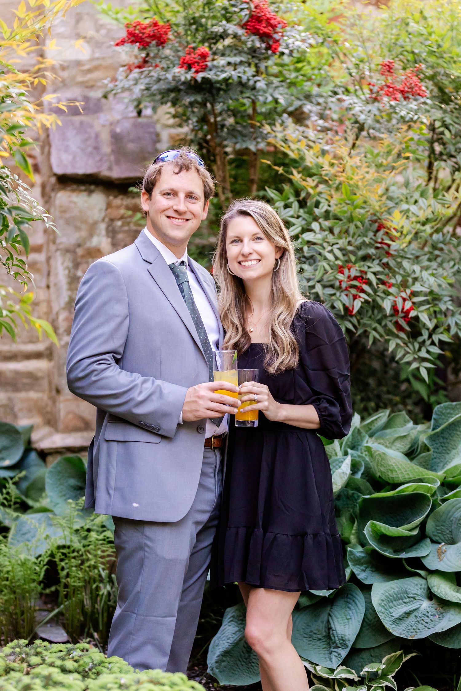 Couple posing in formal attire against red flowering bushes in a garden setting.