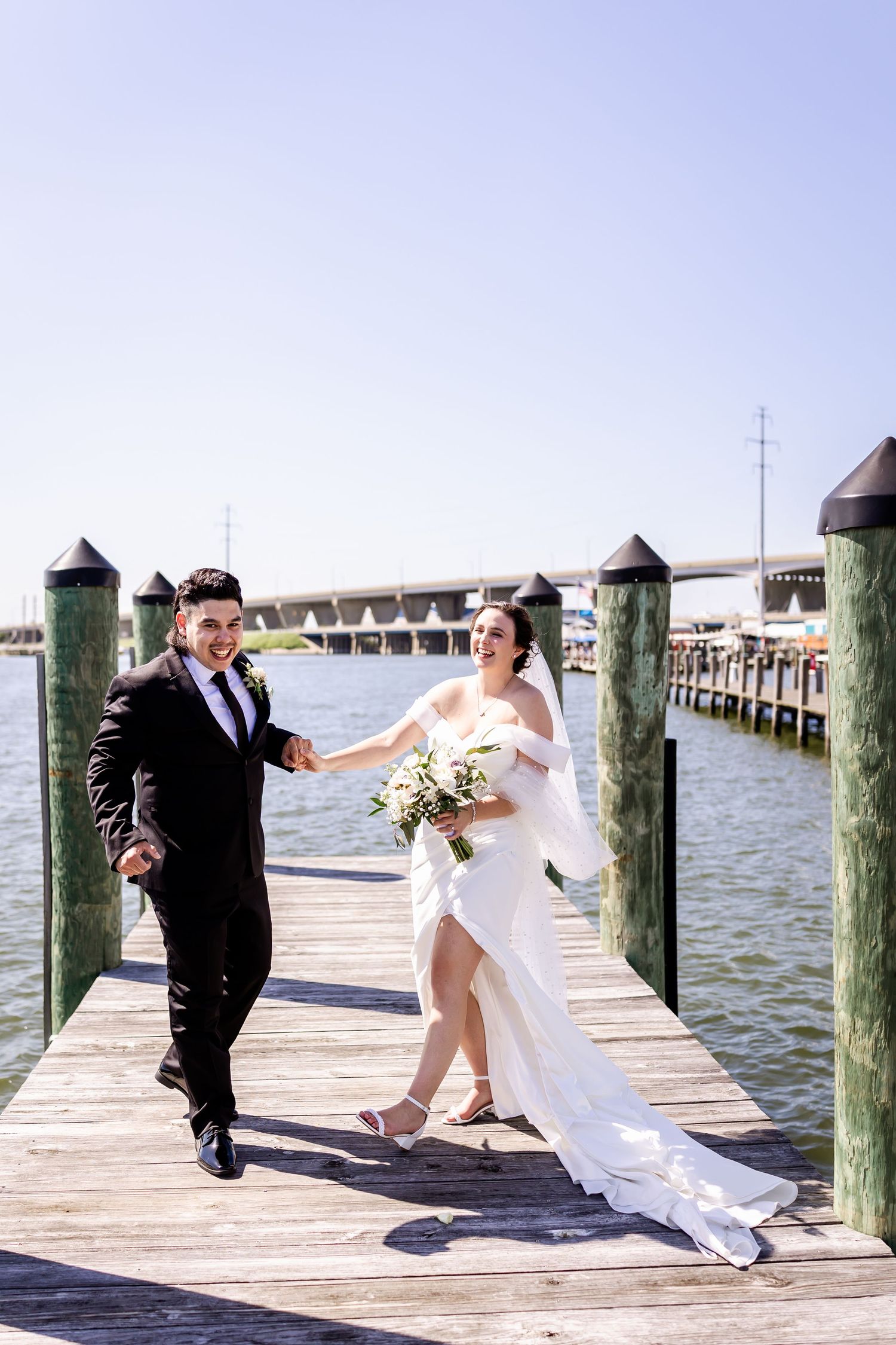 Newlyweds share romantic moment walking along seaside dock with water and boats in background on sunny day.