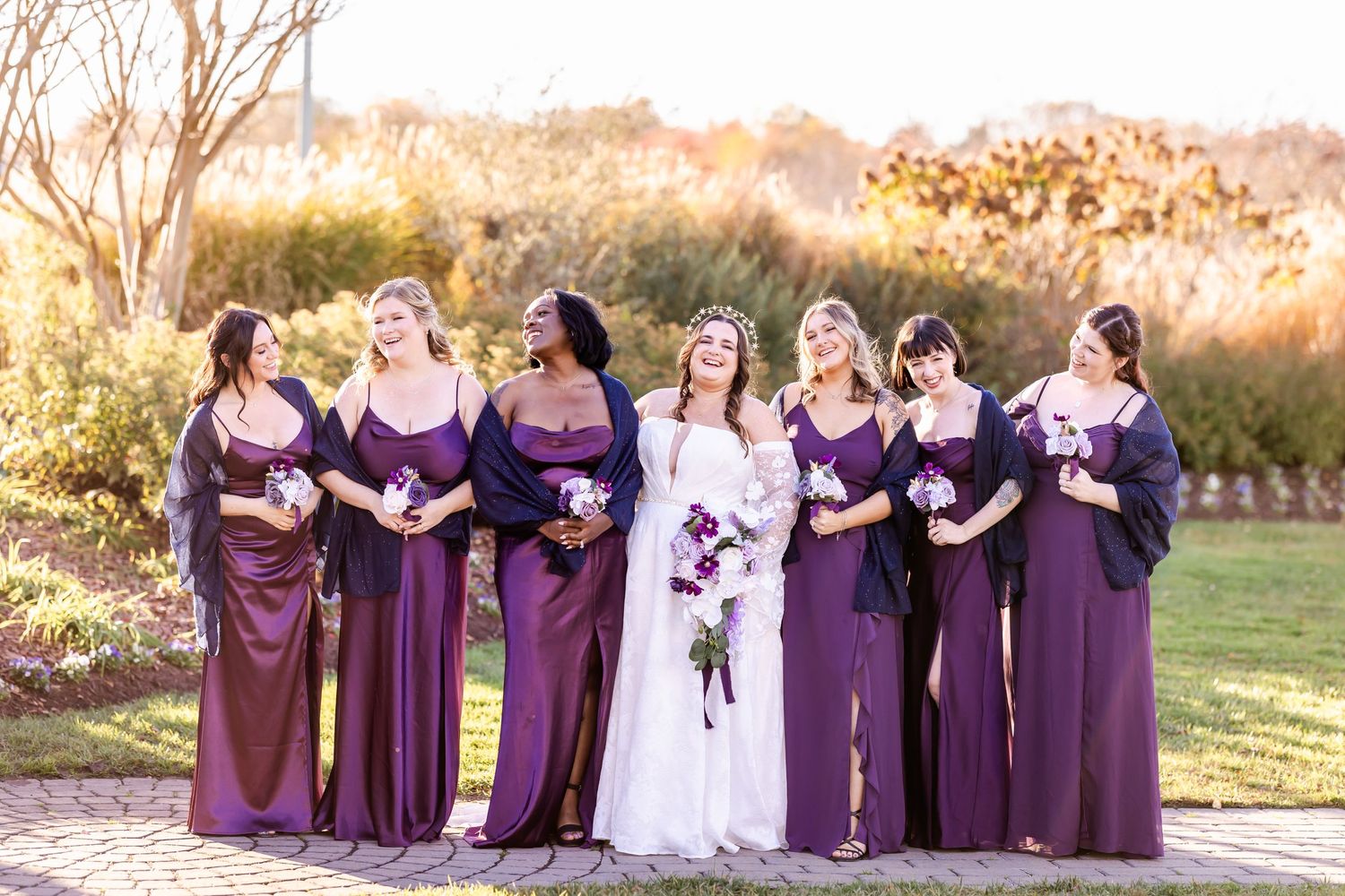 Wedding party in purple dresses poses together on a garden path during golden hour.