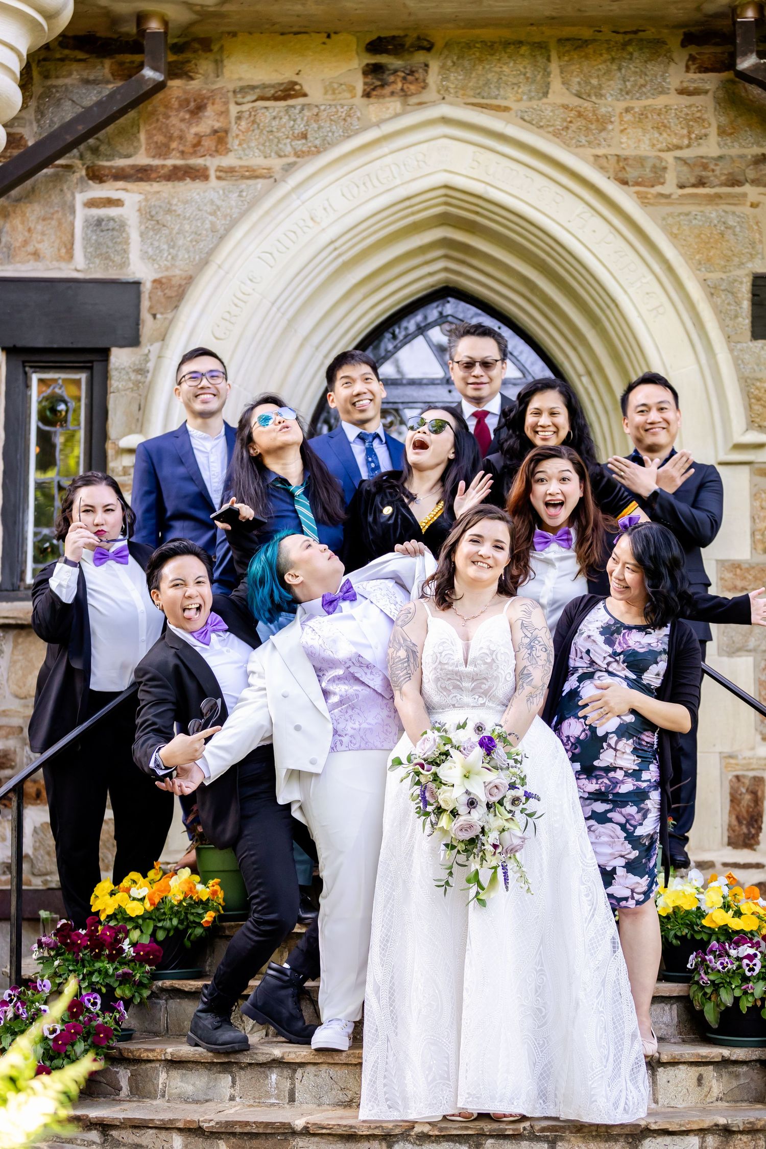 Wedding party poses joyfully on stone church steps with Gothic archway and colorful flowers in foreground.