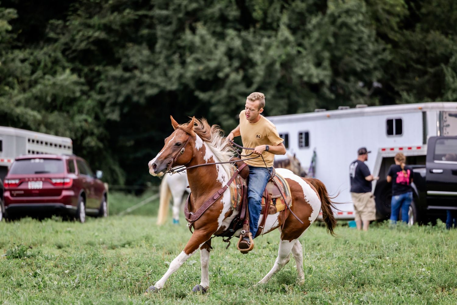 Rider cantering through a grassy field near horse trailers and spectators.