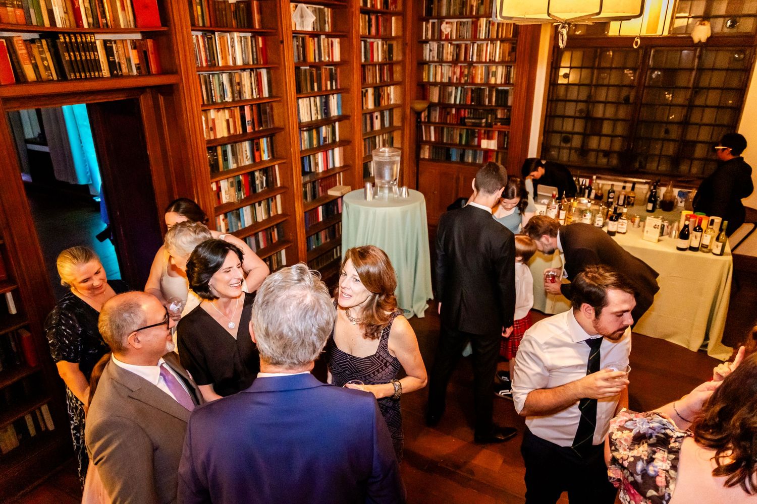 Guests enjoying an evening event in a cozy library setting with floor-to-ceiling bookshelves and warm lighting.