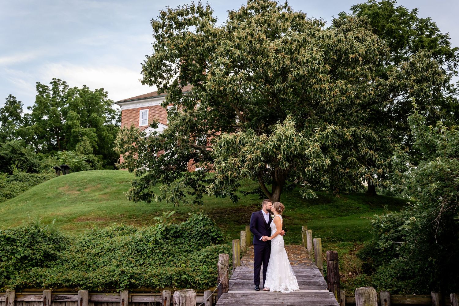 A couple embraces on a stone bridge surrounded by lush greenery and trees at a garden wedding venue.