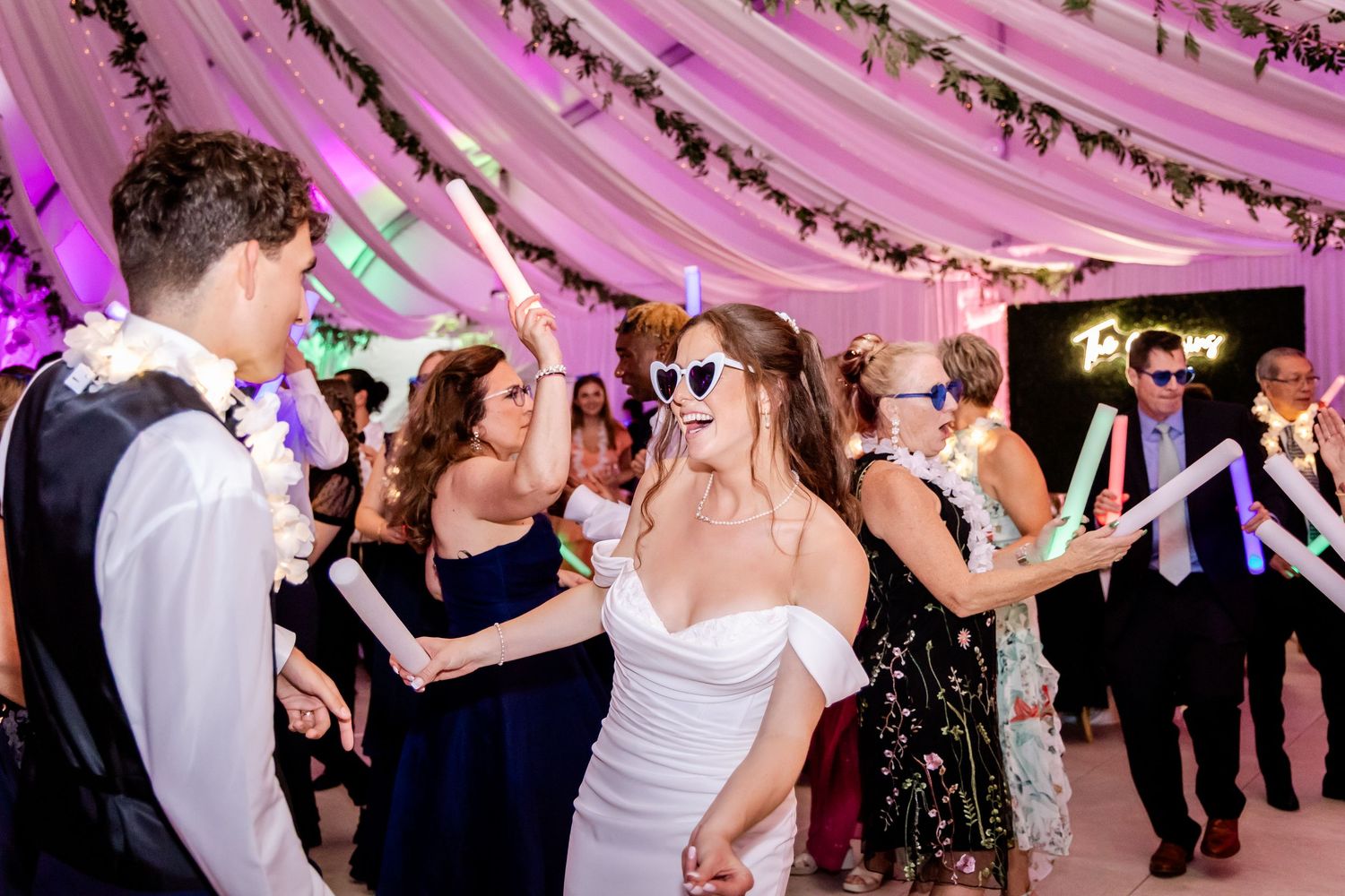 Wedding guests dance and celebrate under pink draped fabric and string lights at a reception.